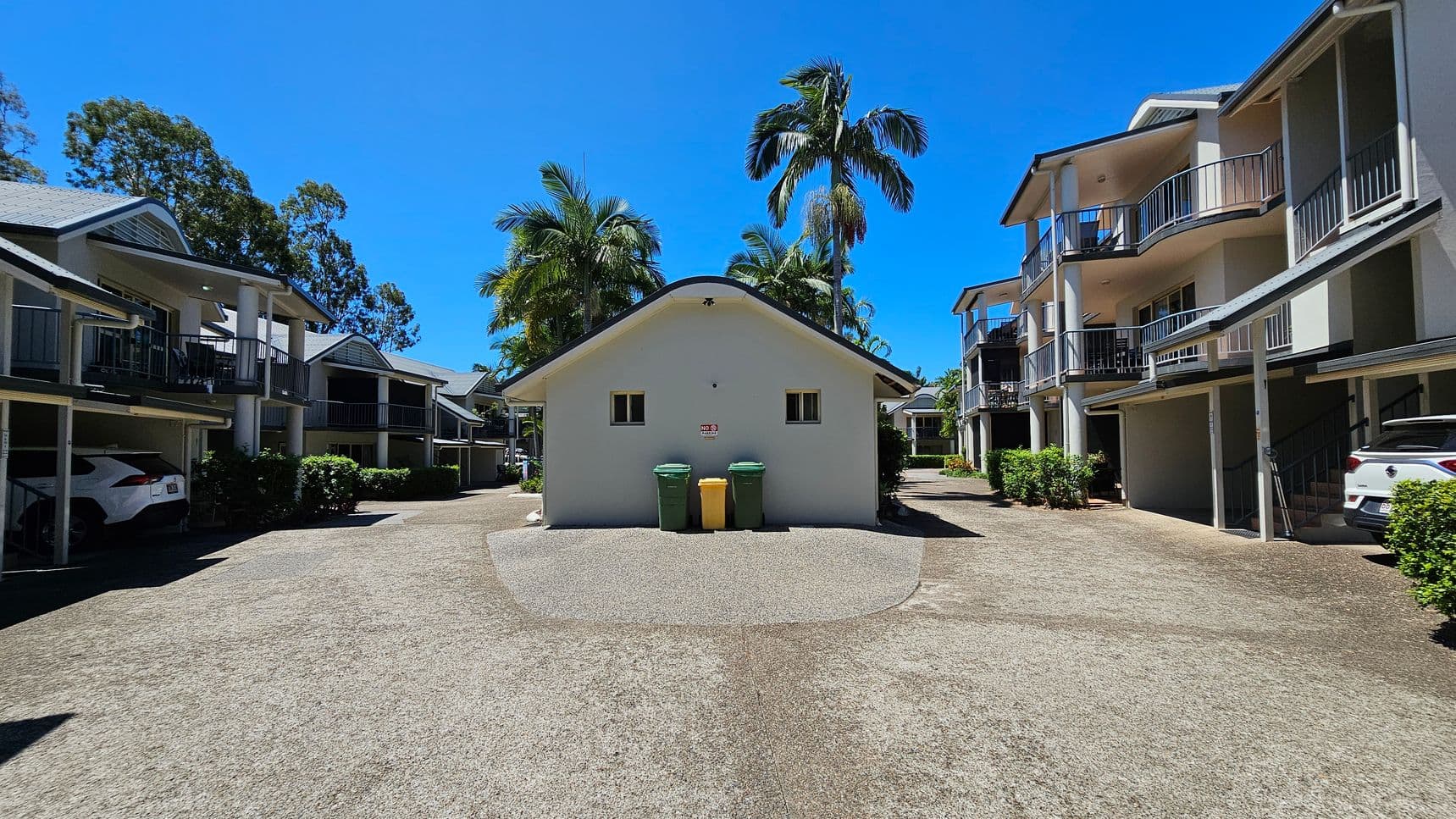 A courtyard with a central building featuring two small windows and three bins in front. On either side, there are two-storey residential buildings with balconies and carports. Cars are parked under the carports. Palm trees and other greenery are visible around the area. The ground is a mix of concrete and gravel.