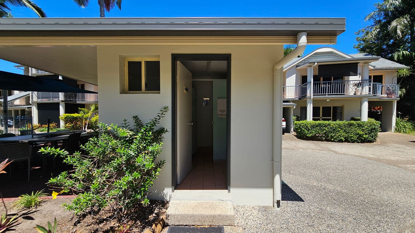 A small, light-coloured building with a narrow entrance in the centre. To the left, there are green shrubs and a shaded outdoor seating area with tables and chairs. On the right, a larger two-storey building with balconies and a hedge in front. The ground is a mix of concrete and gravel, and there are palm trees in the background.