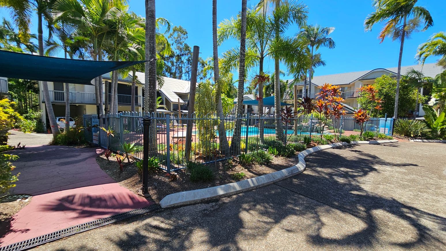 A tropical outdoor setting with a swimming pool surrounded by a metal fence in the centre. Tall palm trees and various plants are scattered around the area. On the left, there is a shaded pathway leading to a building with a parked car. On the right, more plants and a curved concrete barrier are visible. In the background, there are two-storey buildings with balconies. The ground is paved, and the sky is clear.