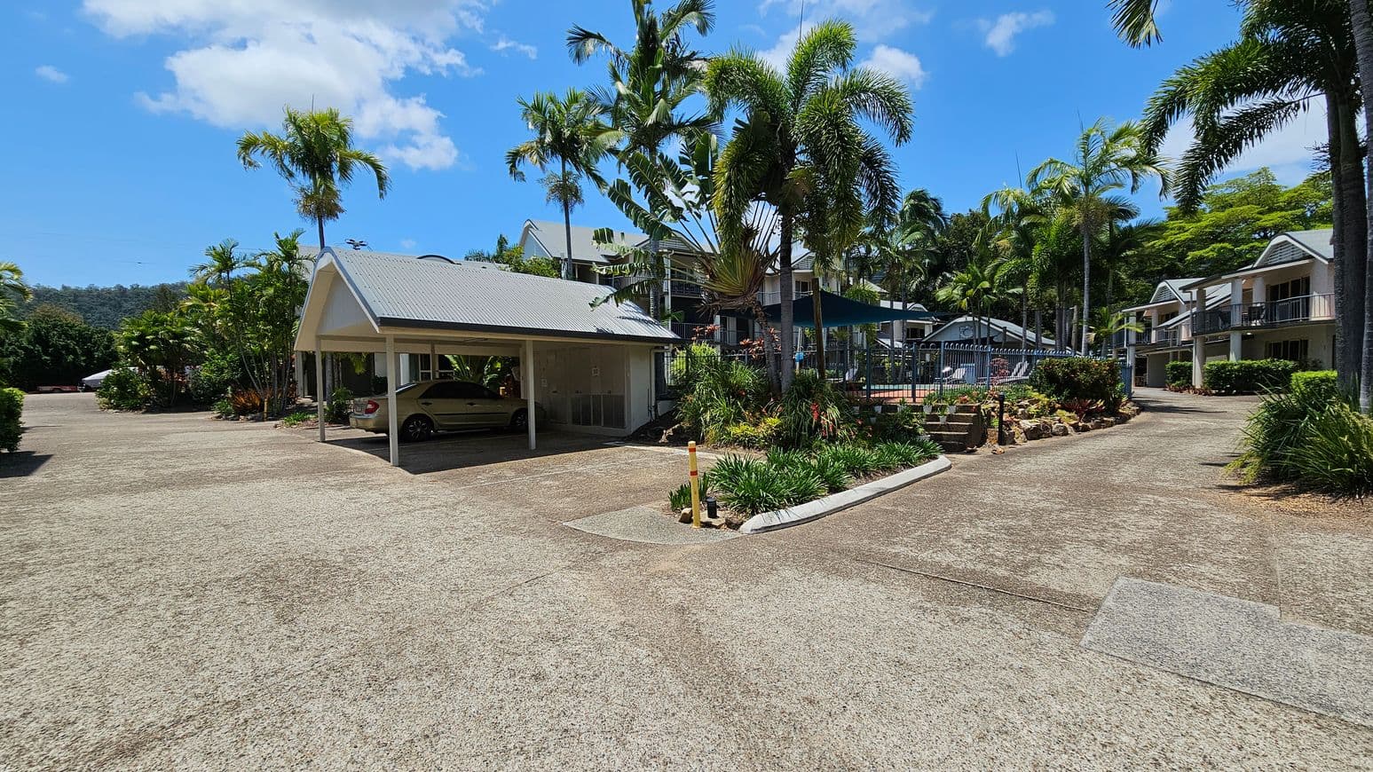 A paved area with a carport on the left, sheltering a parked car. Palm trees and lush greenery surround the area. In the centre, a pathway leads to a fenced pool area with a shade structure. On the right, there are two-storey buildings with balconies, surrounded by more palm trees and shrubs. The sky is clear with a few clouds.