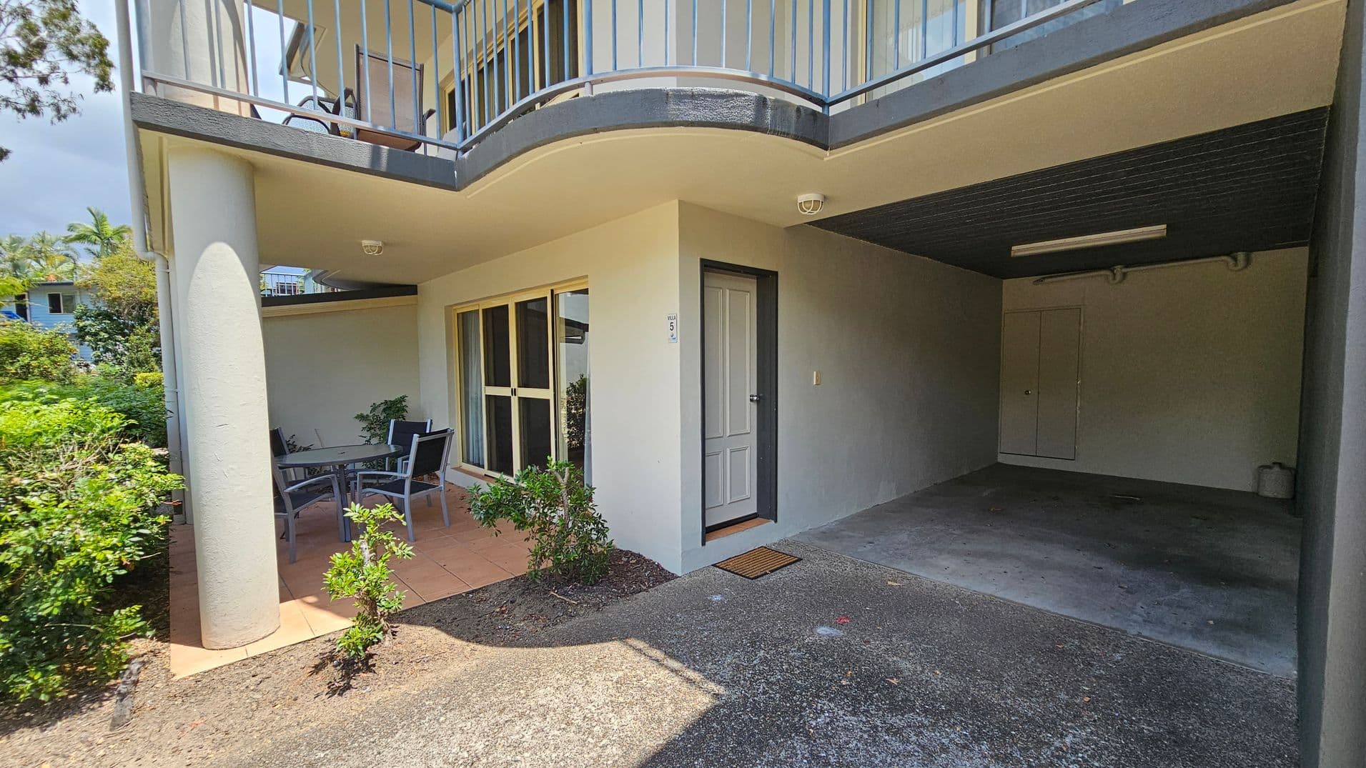 A ground-level outdoor area with a covered patio on the left, featuring a table and chairs. The patio is bordered by a low wall and greenery. A sliding glass door and a regular door are visible on the wall. On the right, there's a covered carport with a concrete floor and a small storage door at the back. The ceiling is flat with a light fixture. Above, a balcony with a metal railing is visible. The ground is a mix of concrete and tiled surfaces.