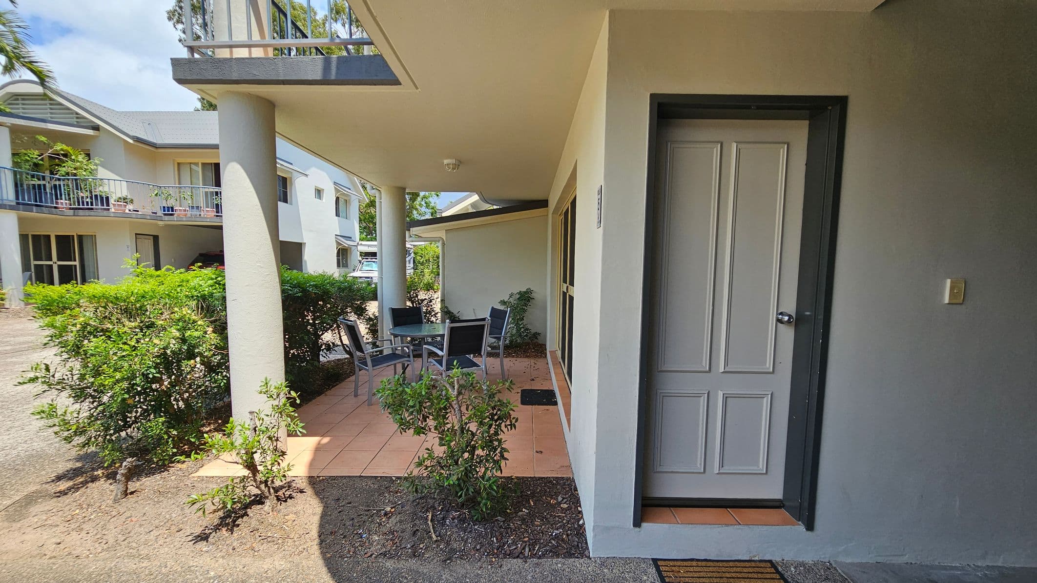 A covered patio area with a tiled floor and a round glass table surrounded by four chairs. To the right, there's a closed door with a doormat in front. The patio is bordered by a low hedge and a few small plants. In the background, there are two-storey buildings with balconies and potted plants. A driveway is visible on the left side. The ceiling of the patio is plain and has a light fixture.