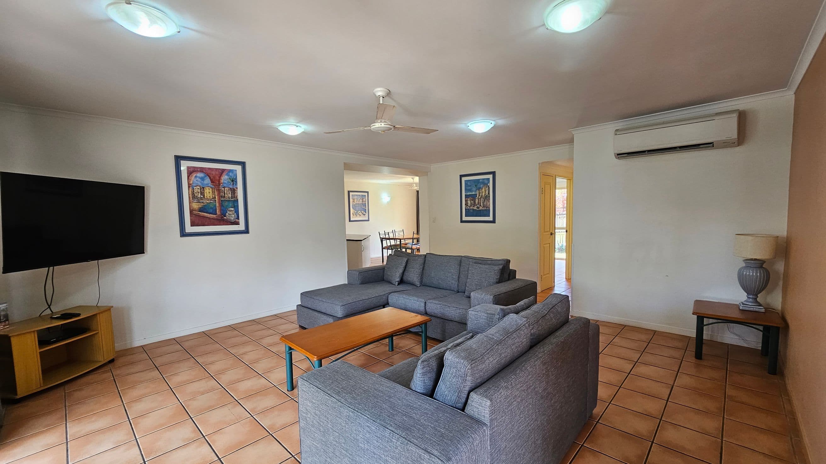 A living room with a tiled floor and a ceiling fan. On the left, a flat-screen TV is mounted on a wooden stand. In the centre, a grey L-shaped sofa faces a wooden coffee table. The right side features a side table with a lamp and an air conditioning unit above. The walls are adorned with framed artwork. In the background, a dining area with a table and chairs is visible through an open doorway.