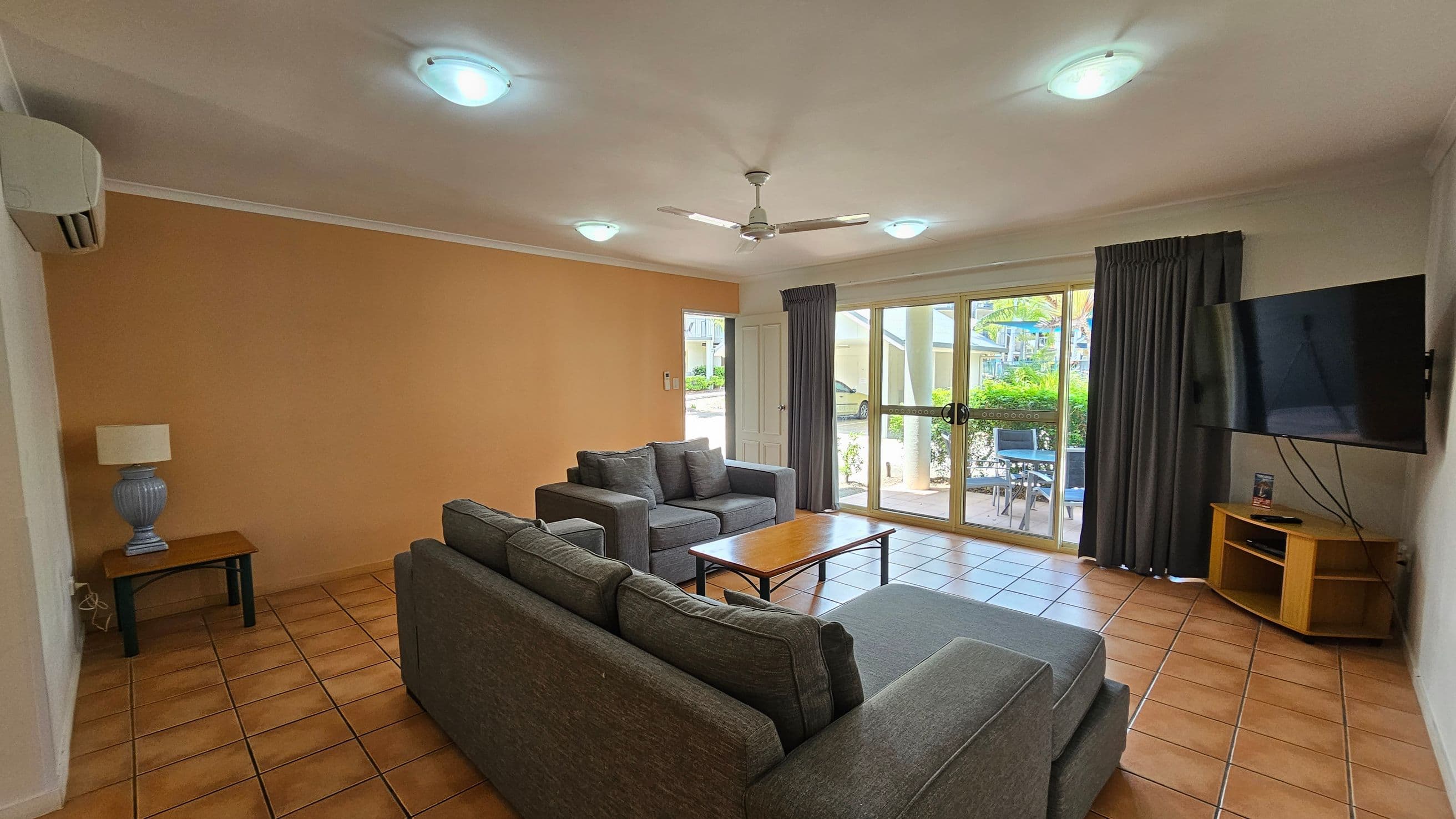 A living room with terracotta tiled flooring and a white ceiling featuring recessed lights and a ceiling fan. On the left, there's a small table with a lamp and an air conditioning unit above. In the centre, two grey sofas face a wooden coffee table. The right side has a wall-mounted TV on a wooden stand. Large glass doors with dark curtains lead to an outdoor area with visible greenery and a table with chairs.