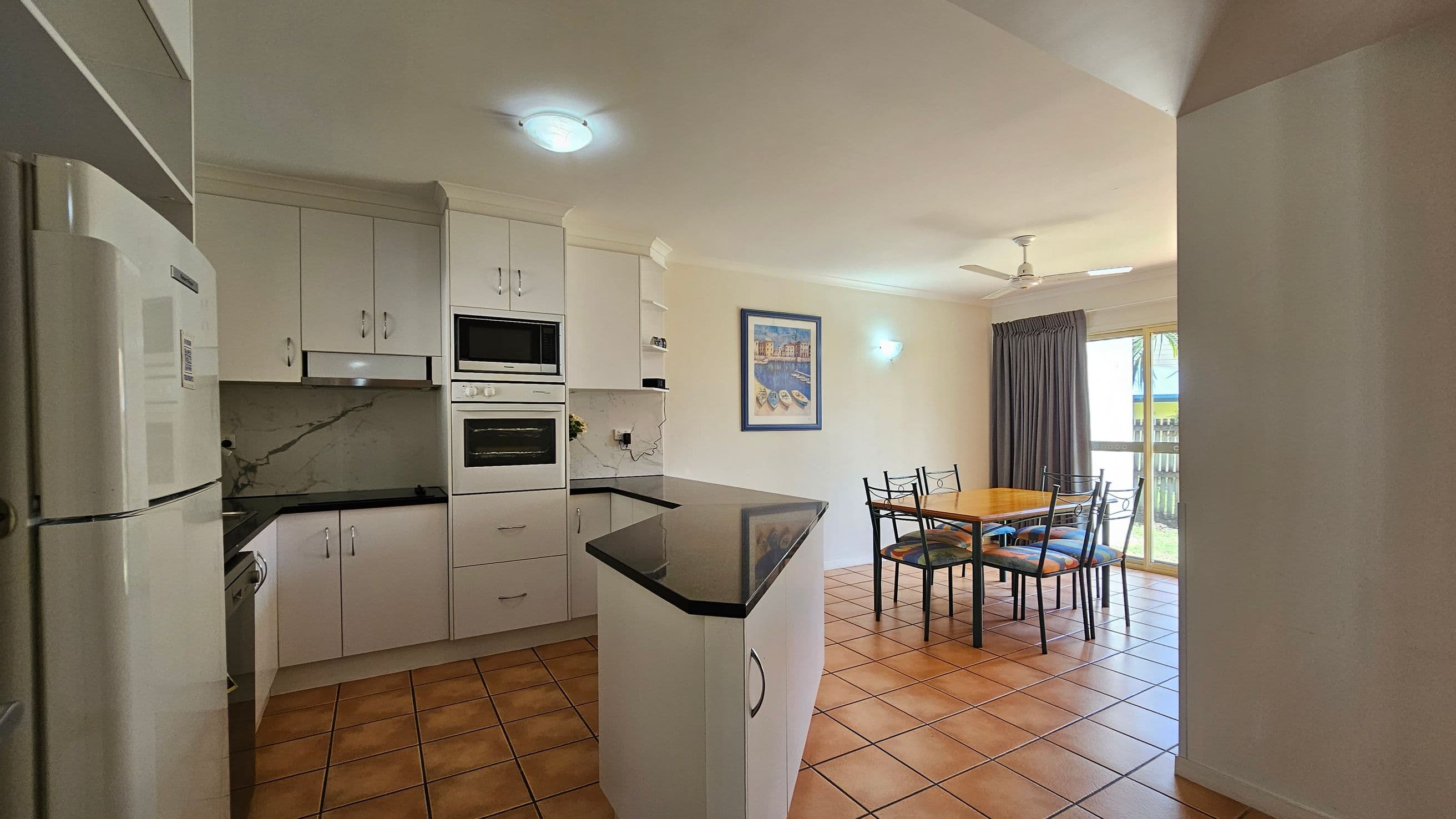 A kitchen and dining area with terracotta floor tiles. On the left, there is a white refrigerator and cabinetry with a built-in oven and microwave. A black countertop extends into the room. In the middle, a dining table with four chairs is set against a wall with a framed picture. On the right, a large window with dark curtains and a ceiling fan above. The ceiling is white with recessed lighting.
