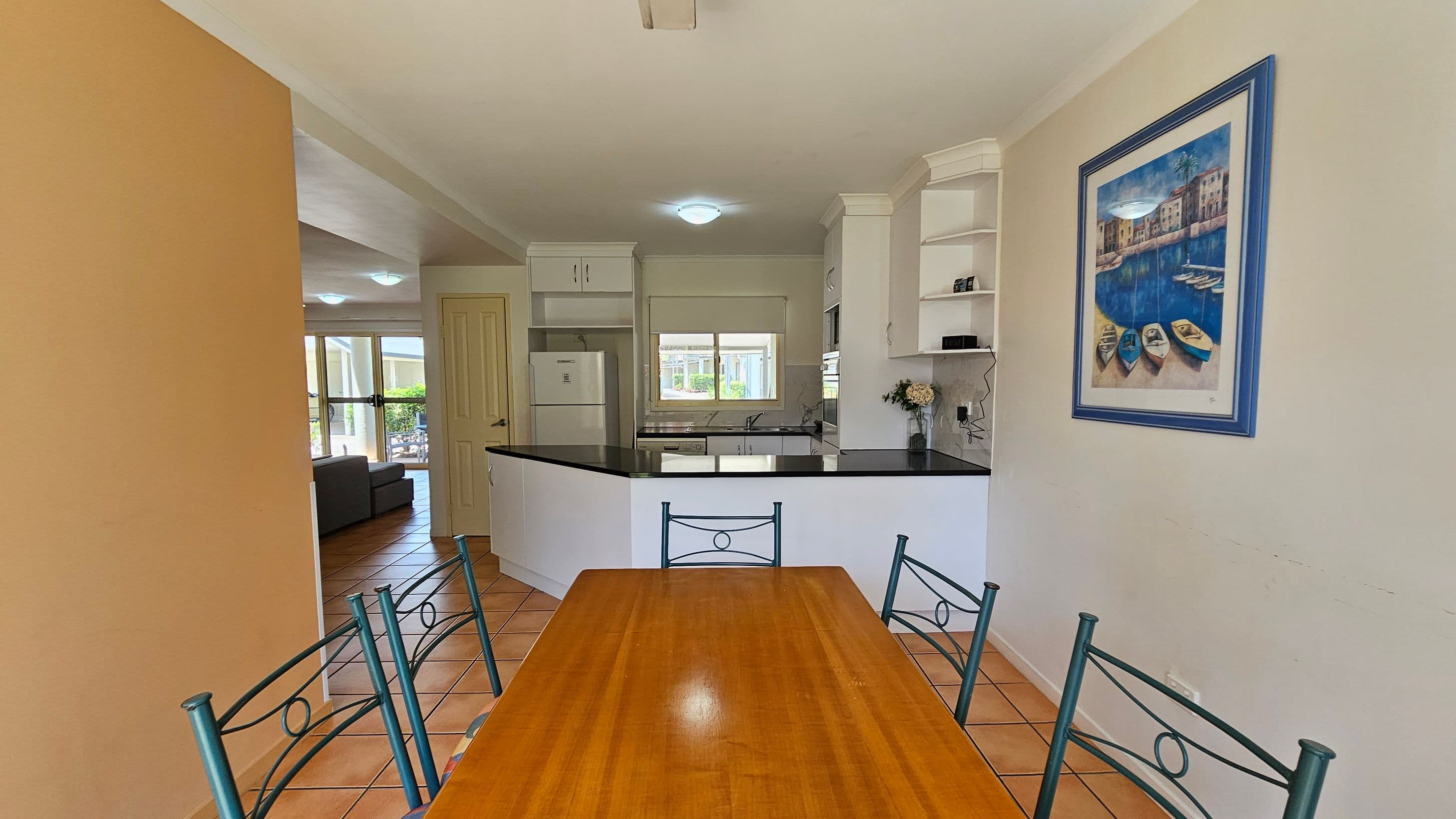 A dining area with a wooden table and metal chairs in the foreground. To the right, a framed painting of boats on water hangs on the wall. In the background, a kitchen with white cabinets, a black countertop, and a window. A refrigerator is visible to the left of the kitchen. Beyond the kitchen, a living area with a grey sofa and large windows can be seen. The floor is tiled with a terracotta colour.