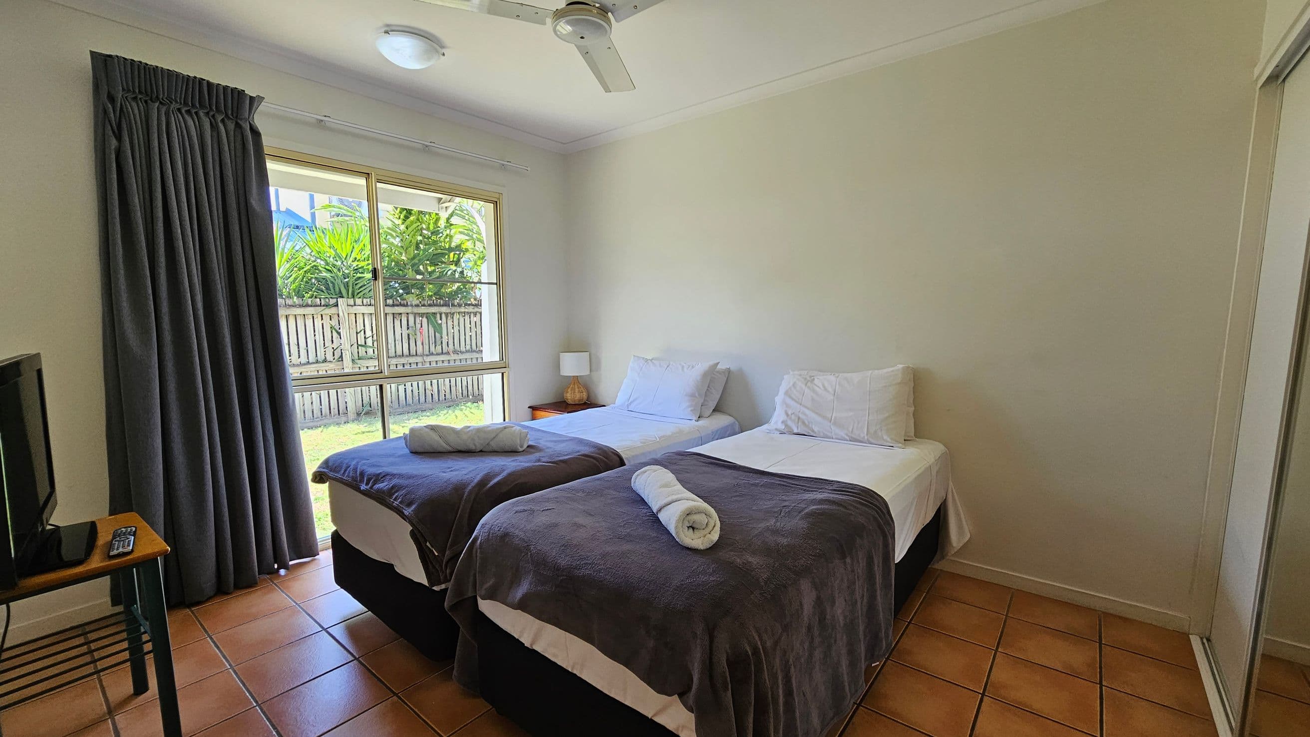 A bedroom with two single beds in the centre, each with white sheets, pillows, and a rolled towel on top of dark blankets. A small bedside table with a lamp is between the beds. On the left, a window with closed dark curtains overlooks a garden with a wooden fence and greenery. A TV sits on a small table with a remote control. The floor is tiled in a terracotta colour, and a ceiling fan is visible above. A mirrored wardrobe is on the right.