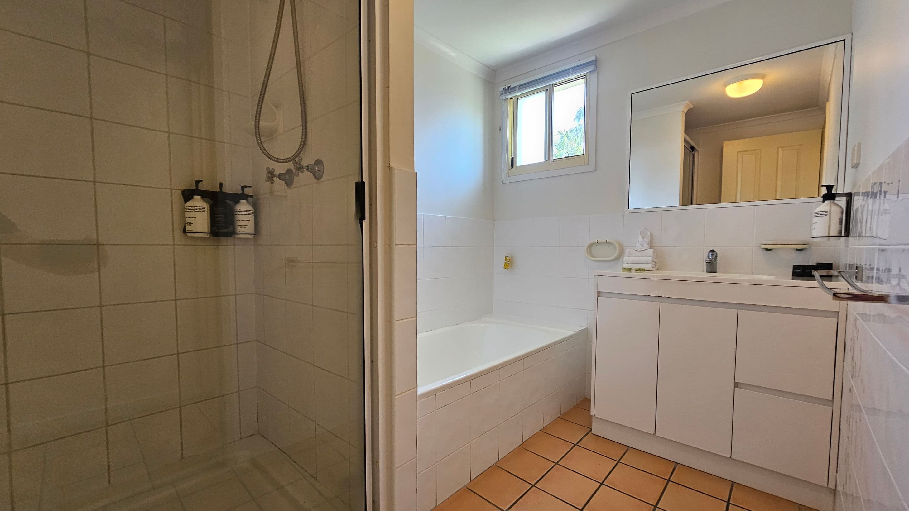 A bathroom with a tiled floor and white walls. On the left, there's a glass-enclosed shower with mounted soap dispensers. In the centre, a white bathtub is positioned beneath a window with a blind. On the right, a white vanity unit with a large mirror above it holds a soap dispenser and a small decorative item. The ceiling light is visible in the mirror's reflection.