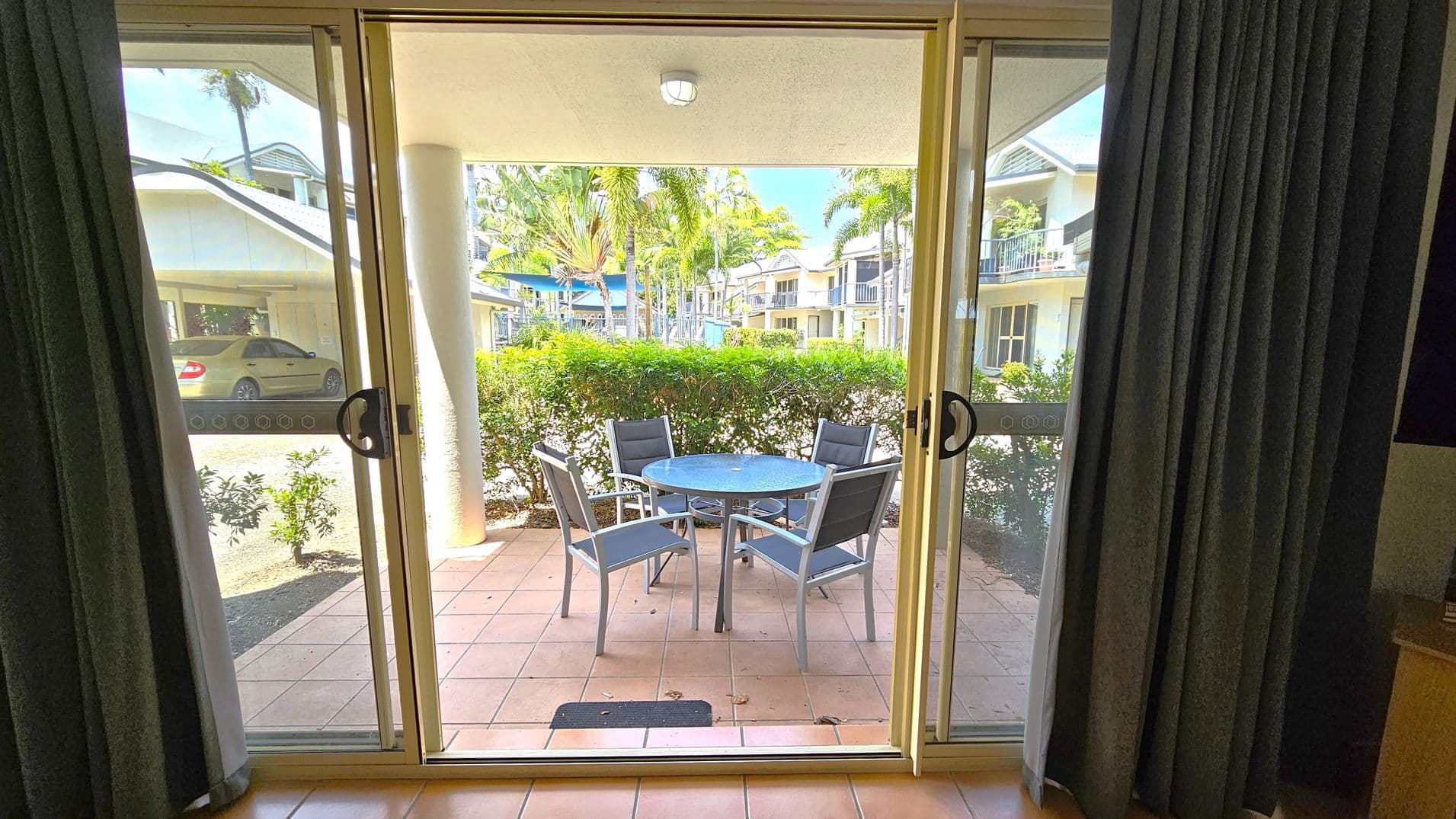 Sliding glass doors open to a patio with terracotta tiles. Outside, there's a round glass table with four grey chairs. A hedge borders the patio, and beyond it, palm trees and a view of a residential area with white buildings and balconies. A yellow car is parked on the left. Dark curtains frame the doors from the inside.