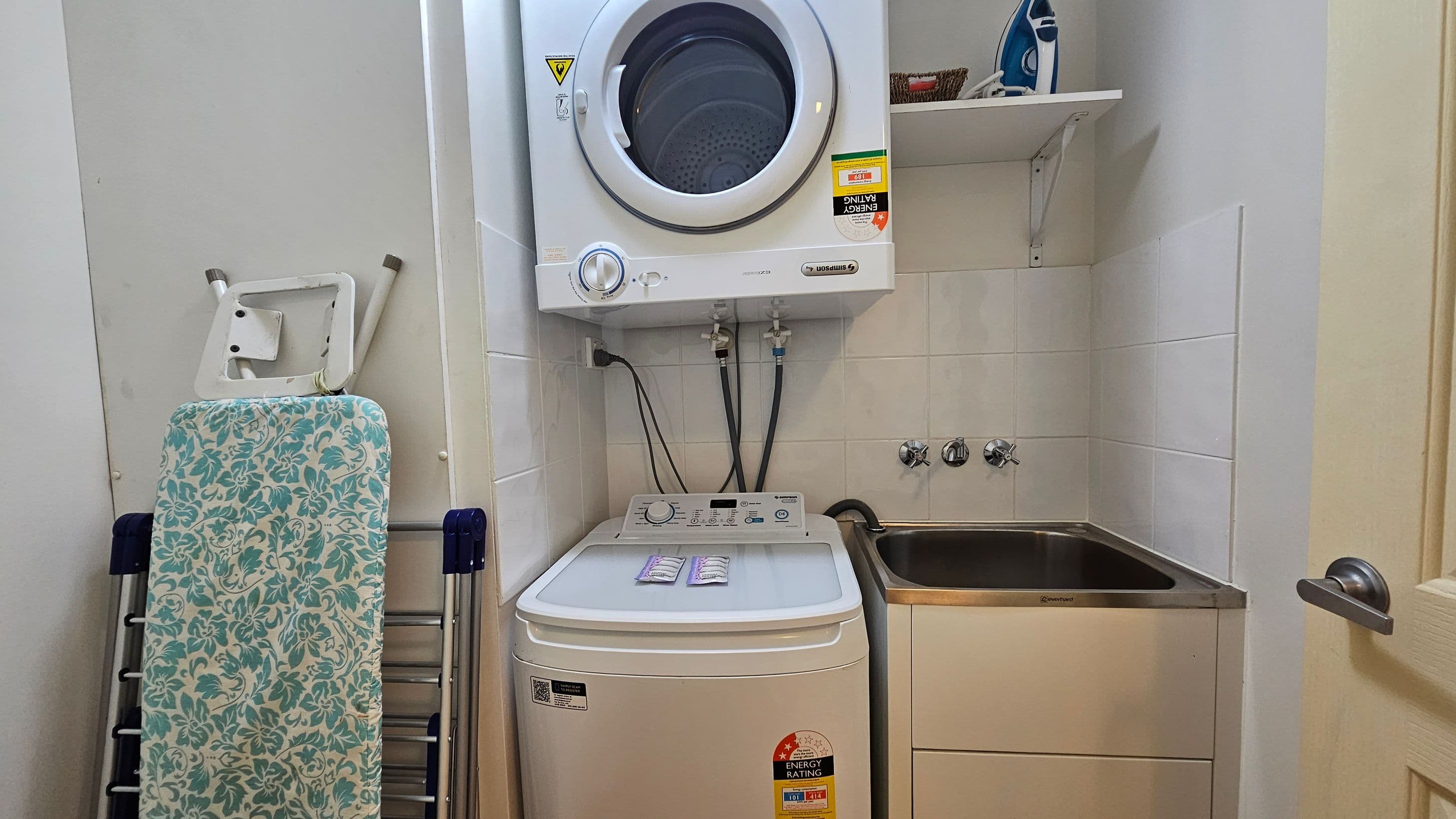 A compact laundry area with a top-loading washing machine and a front-loading dryer stacked above it. To the left, there's a folded ironing board with a floral cover. On the right, a stainless steel sink is set into a cabinet. Above the sink, a shelf holds an iron and a small basket. The walls are tiled, and a door with a silver handle is partially visible on the far right.