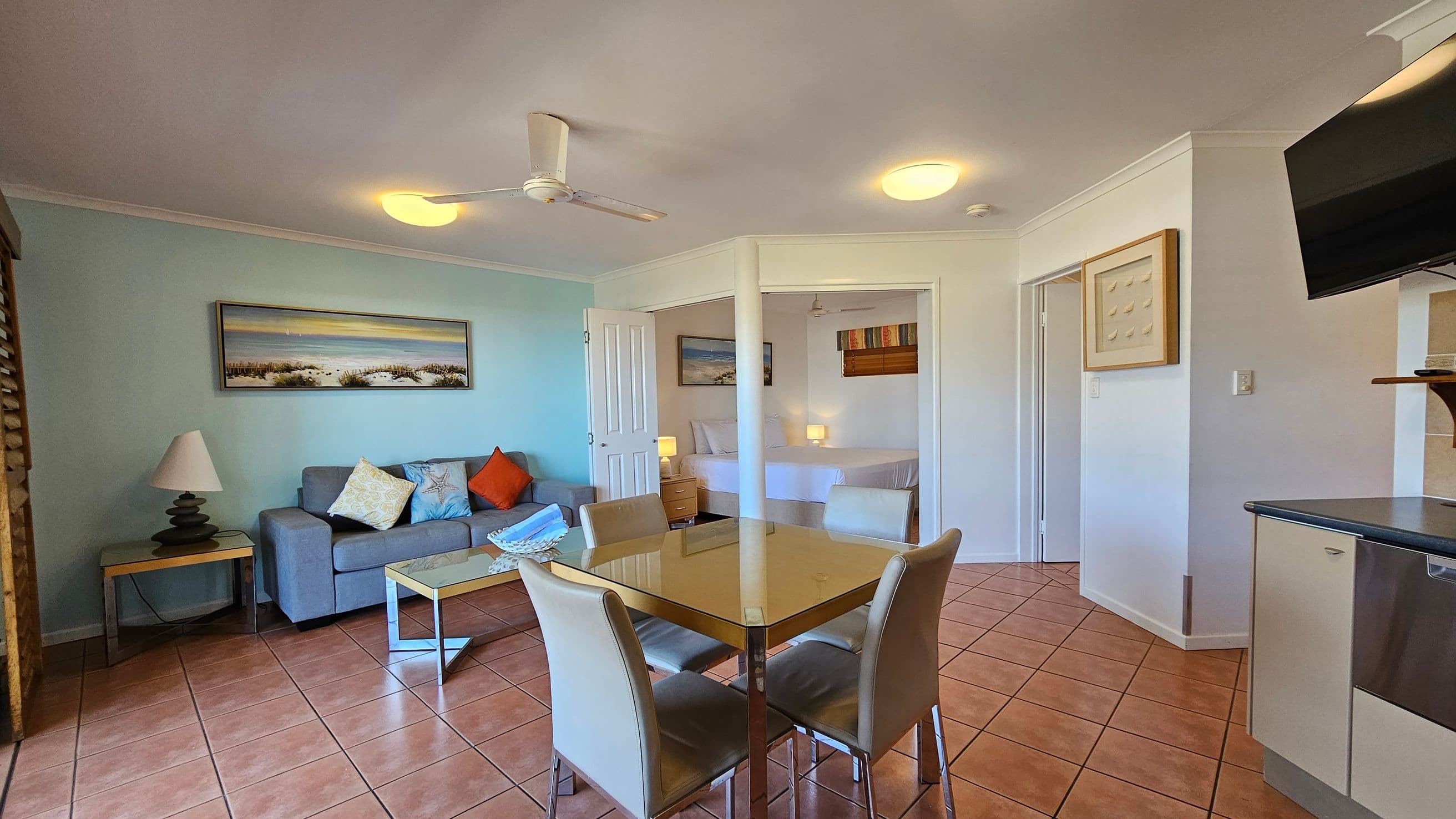 A living room with terracotta tiled flooring and a ceiling fan. On the left, there are wooden shutters leading to a balcony with a view of green hills. In the centre, a glass dining table with four chairs is placed. On the right, a grey sofa with colourful cushions sits against a light blue wall, above which hangs a landscape painting. A glass coffee table is in front of the sofa. A small wooden cabinet with a lamp is near a white door.
