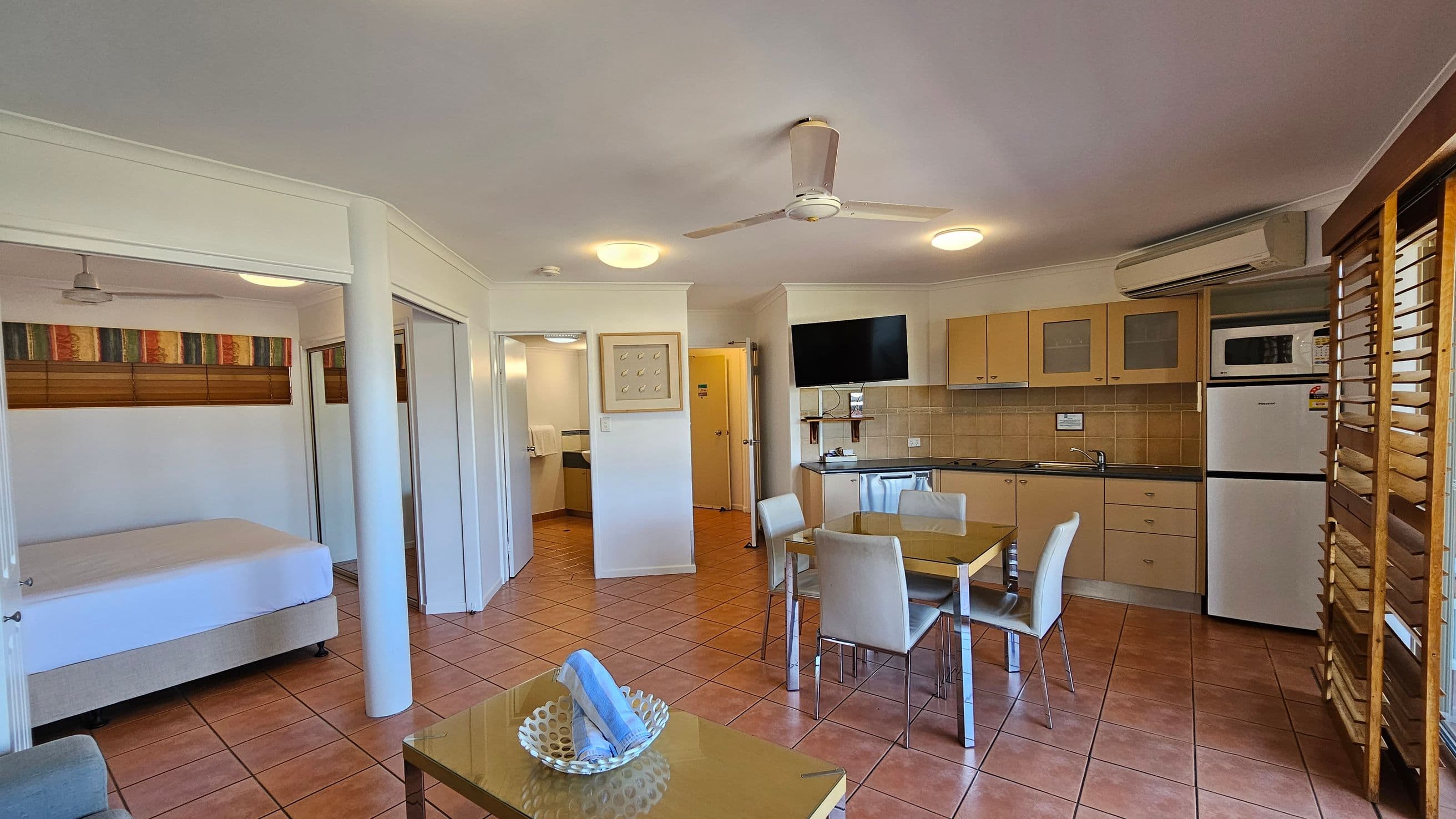 A room with terracotta tiled flooring and a white ceiling with a ceiling fan. On the left, there's a bed with a colourful headboard and a ceiling fan above. In the centre, a dining table with four chairs is placed near a kitchenette with light wood cabinets, a microwave, and a fridge. A TV is mounted on the wall above the kitchen counter. On the right, wooden louvres cover the windows. A small table with a decorative bowl and rolled towels is in the foreground.