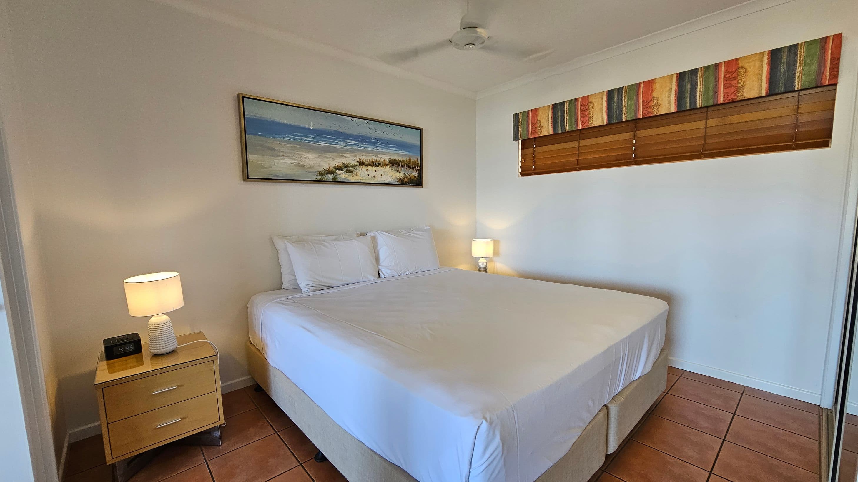 A bedroom with a large bed in the centre, covered in white linens and pillows. On the left, a wooden bedside table holds a lamp and a digital clock. Above the bed, a framed beach scene painting hangs on the wall. On the right, a window with wooden blinds and a colourful valance. The floor is tiled in a terracotta colour, and a ceiling fan is visible above.