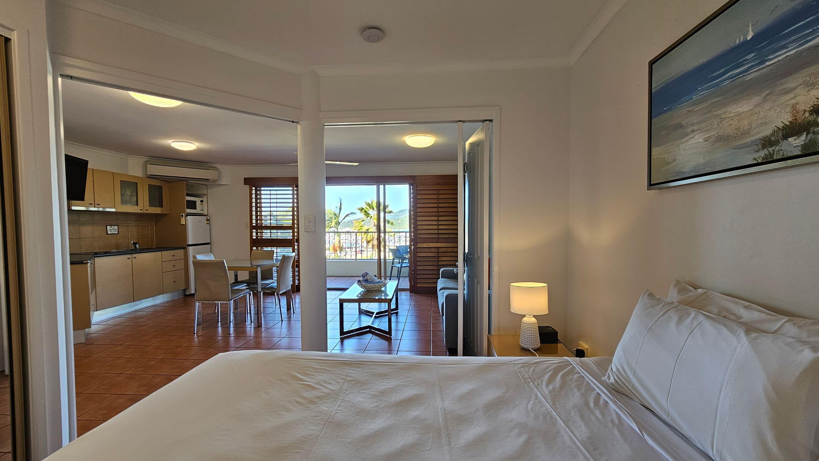 A bedroom with a neatly made bed and a bedside table with a lamp on the right. A framed picture hangs above the bed. Through an open doorway, a living area is visible with a kitchen on the left, featuring light wood cabinets and a small dining table with chairs. Beyond, a sitting area with a sofa and coffee table is seen, leading to a balcony with a view of palm trees. The floor is tiled, and the ceiling has recessed lighting.