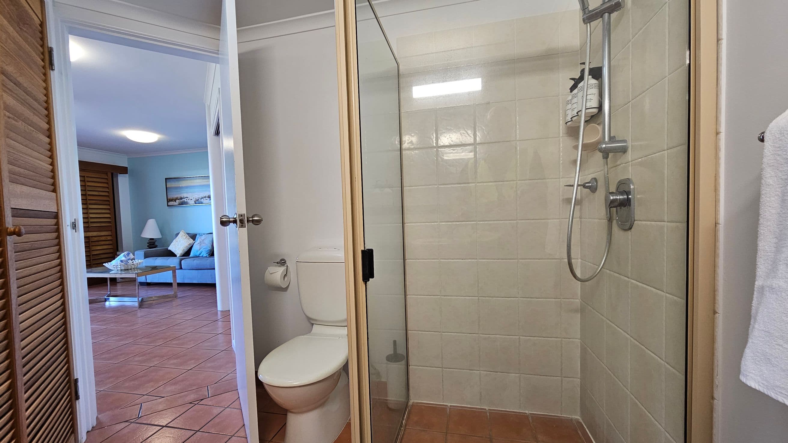 A bathroom with a glass shower enclosure on the right, featuring beige tiles and a silver showerhead. A white toilet is next to the shower, with a toilet roll holder on the wall. The door is open, leading to a room with terracotta floor tiles. In the adjacent room, a blue sofa, a lamp, and a coffee table are visible. A painting hangs on the wall, and wooden shutters cover a window.