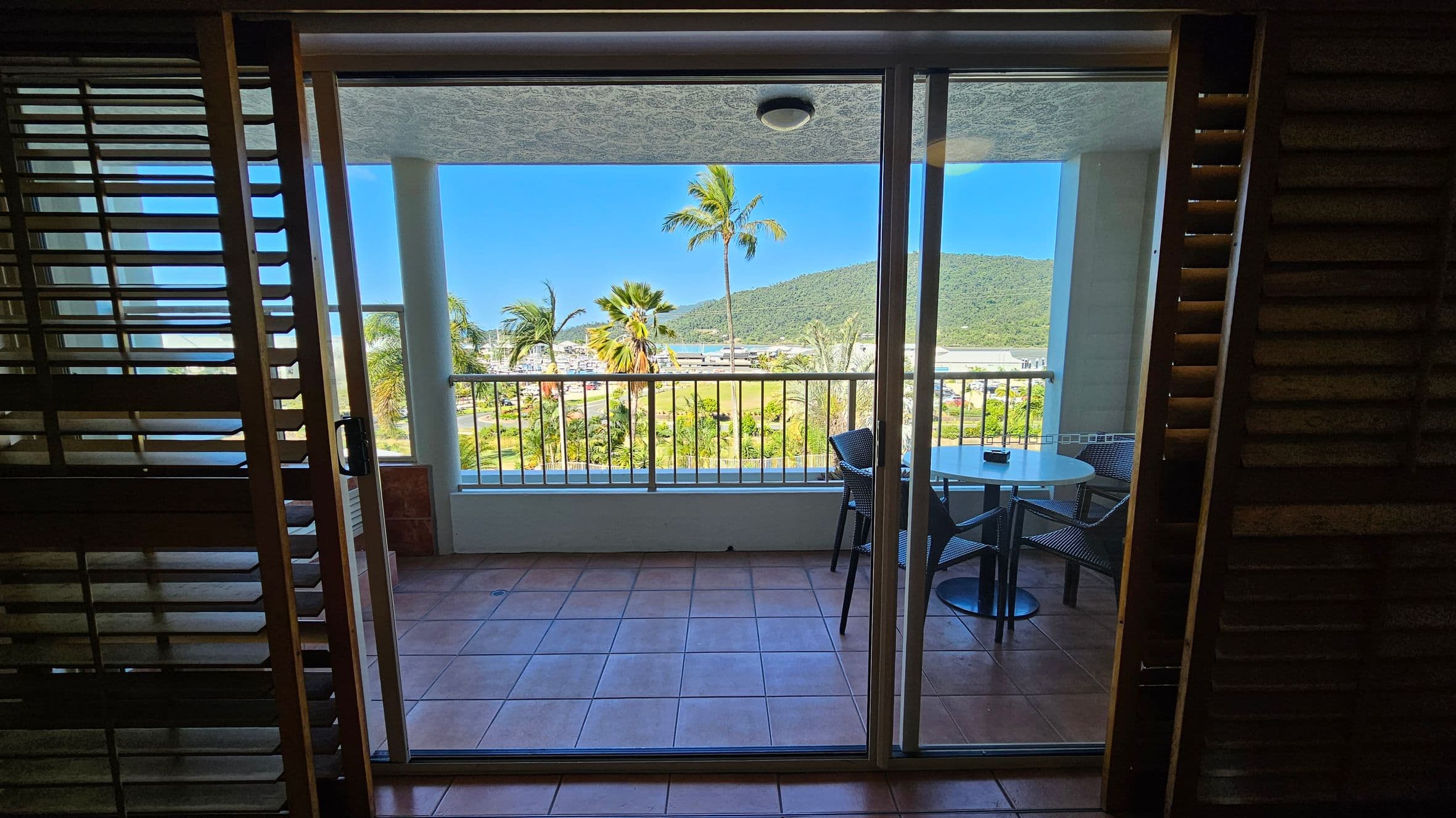 A view through a glass sliding door leading to a balcony with terracotta tiles. On the balcony, there's a round table with two chairs. Beyond the railing, palm trees and lush greenery are visible, with a hill in the background. The ceiling of the balcony is white with a light fixture. Wooden shutters are partially open on either side of the door.