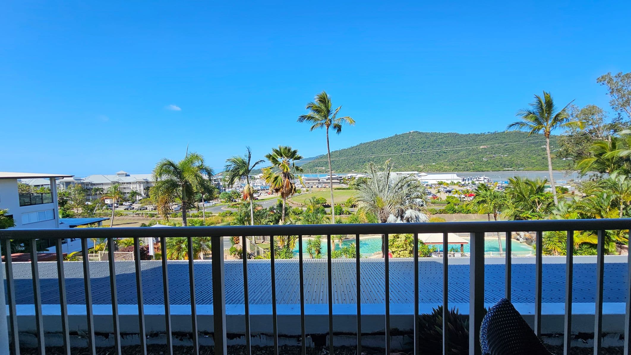 A view from a balcony with a railing in the foreground. Below, there's a corrugated metal roof and a swimming pool surrounded by lush greenery and palm trees. In the background, a small town with buildings and parked cars is visible. Beyond the town, a green hill rises under a clear blue sky.