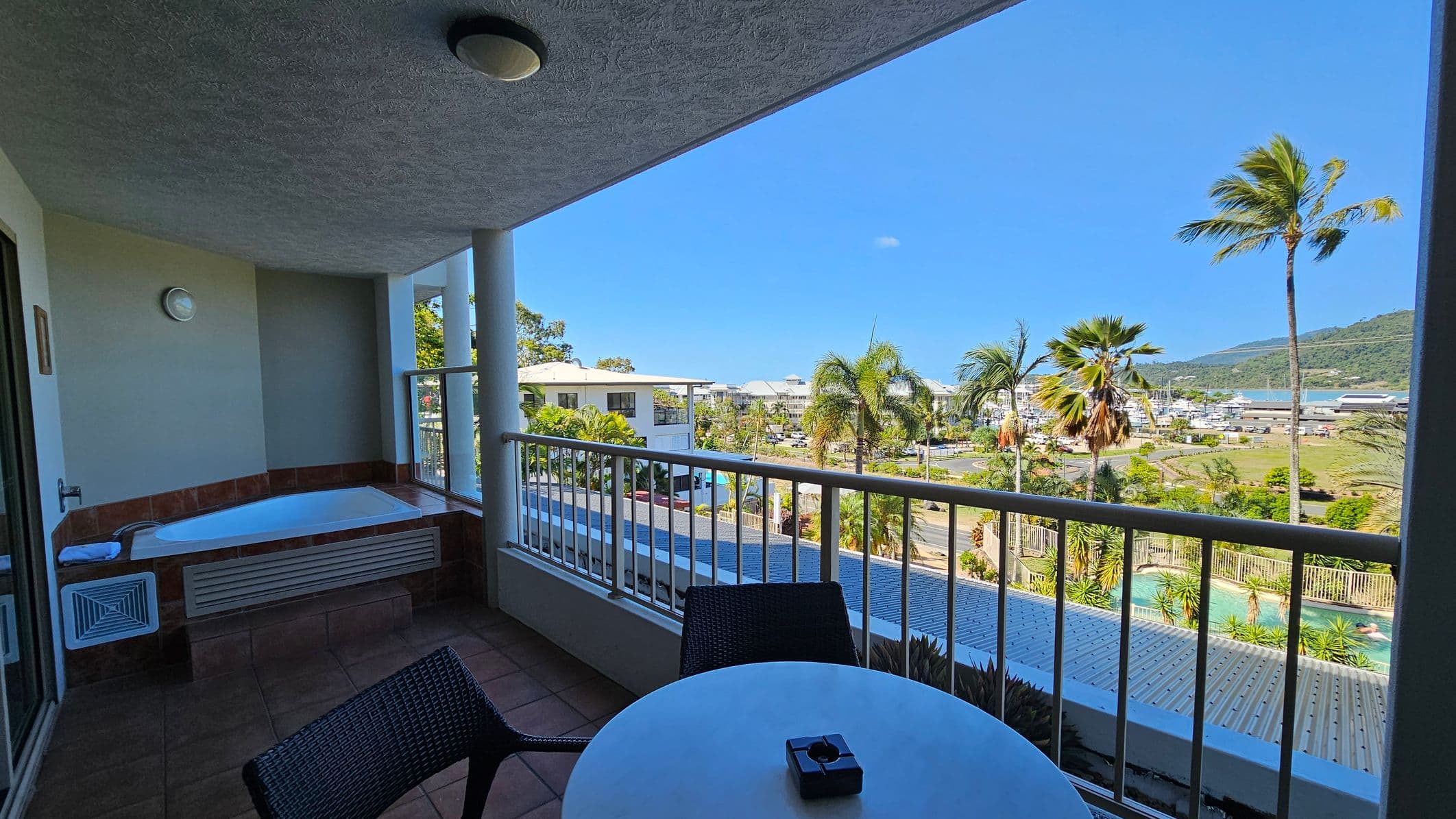 A balcony with a tiled floor features a round table and two chairs. On the left, there's a built-in bathtub with a towel. The railing overlooks a view of palm trees, a swimming pool, and a marina in the distance. The ceiling has a light fixture. The background includes buildings and a hillside.