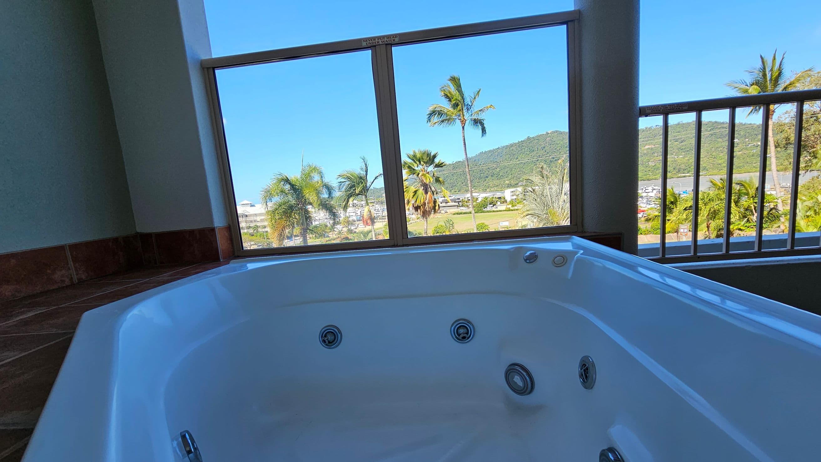 A white jacuzzi tub is in the foreground, positioned indoors near large windows. The windows offer a view of palm trees, a grassy area, and a distant hill. The floor is tiled with dark tiles, and the ceiling is not visible. The scene outside is bright and sunny, with clear blue skies.