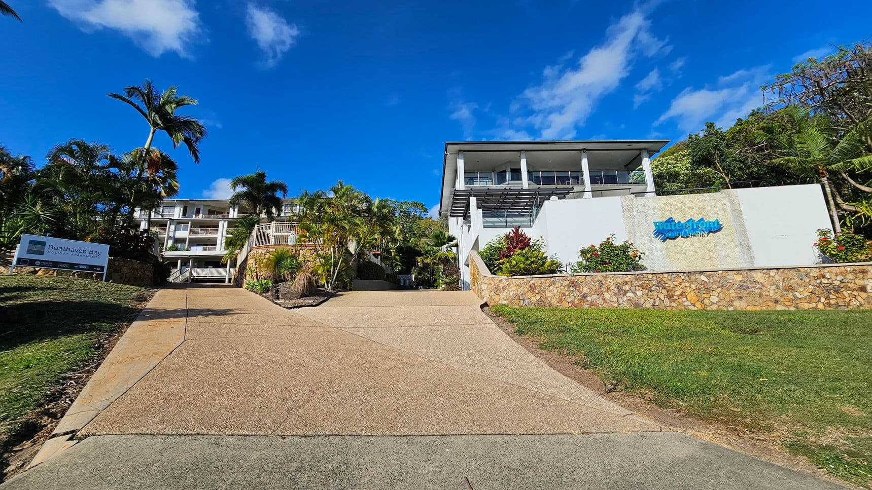 A paved driveway leads up to a multi-storey building surrounded by lush palm trees and tropical plants. On the left, a sign reads "Boathaven Bay Holiday Apartments." The building features balconies and is set against a backdrop of clear blue sky. The foreground includes a grassy slope with scattered rocks and a variety of greenery.