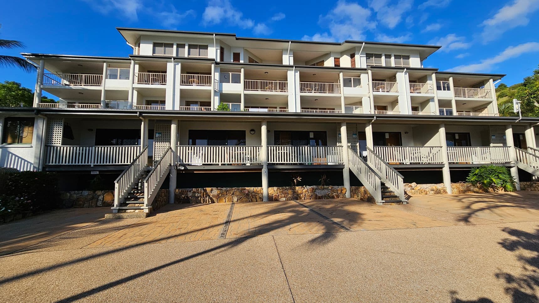 A multi-storey building with a series of balconies and white railings. The structure has a light green facade and is elevated on stone foundations. Several staircases lead up to the main entrance, which is shaded by a roof supported by columns. The foreground features a paved area with shadows of palm trees. The sky is blue with some clouds.