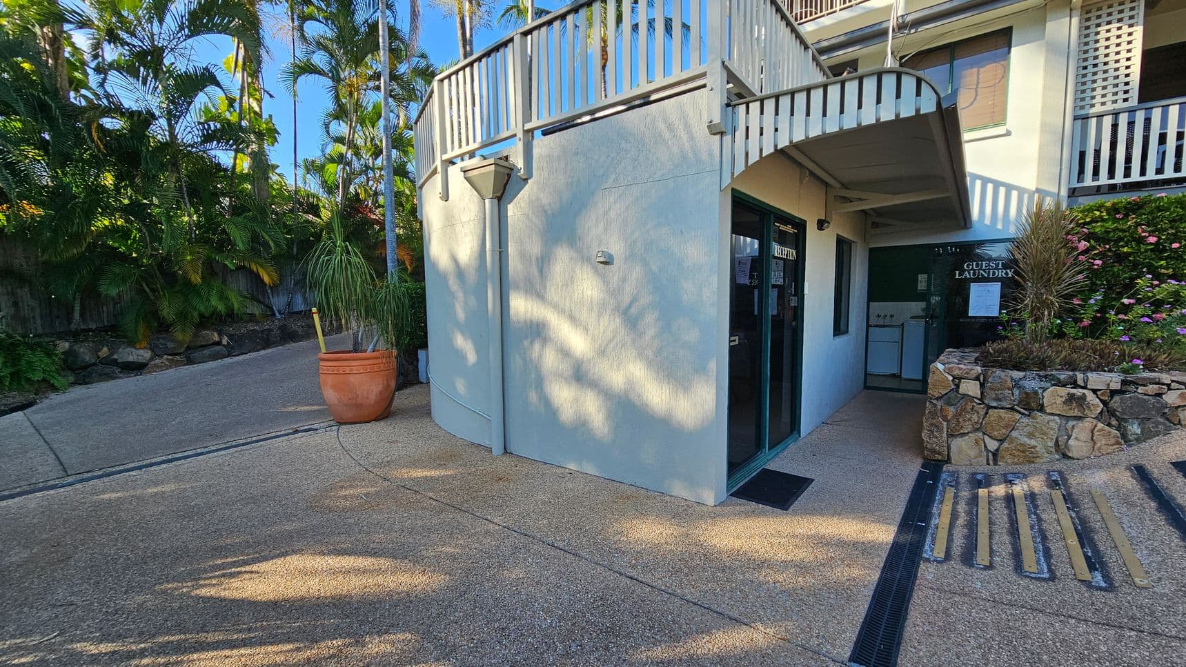 A curved building corner with a glass door labelled "Reception" on the right. Next to it, a sign reads "Guest Laundry" above a stone planter with pink flowers. A large terracotta pot with a tall plant is on the left, near a sloped path lined with lush green palm trees. The ground is textured concrete, and a metal railing runs along the top of the building.