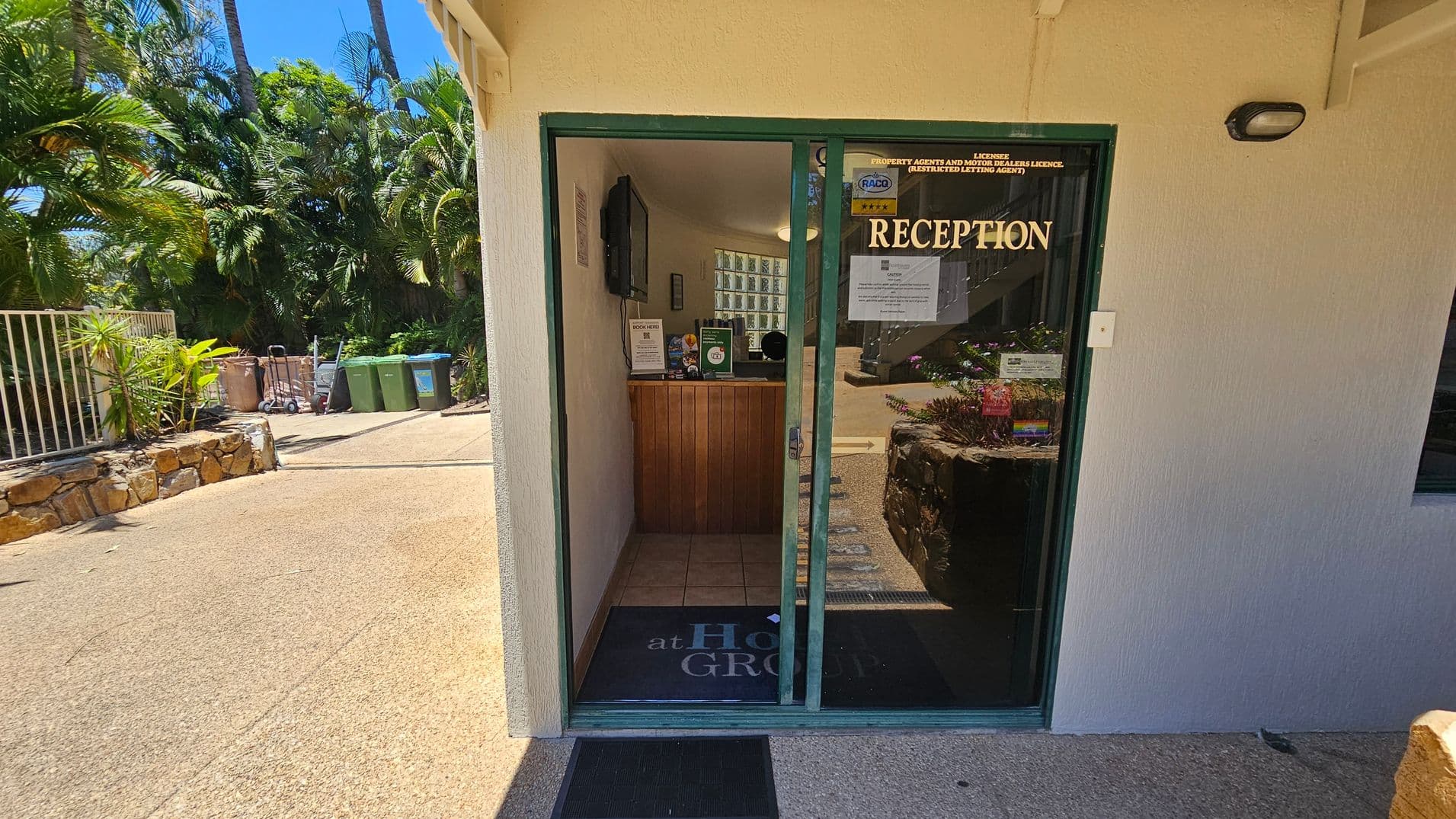 A small reception area with a glass door labelled "RECEPTION" in gold letters. Inside, there's a wooden counter with brochures and a computer screen. The floor is tiled. Outside, to the left, there are green and brown bins, a trolley, and lush greenery with palm trees. A stone wall lines the path leading to the entrance. The building exterior is light-coloured with a textured finish.