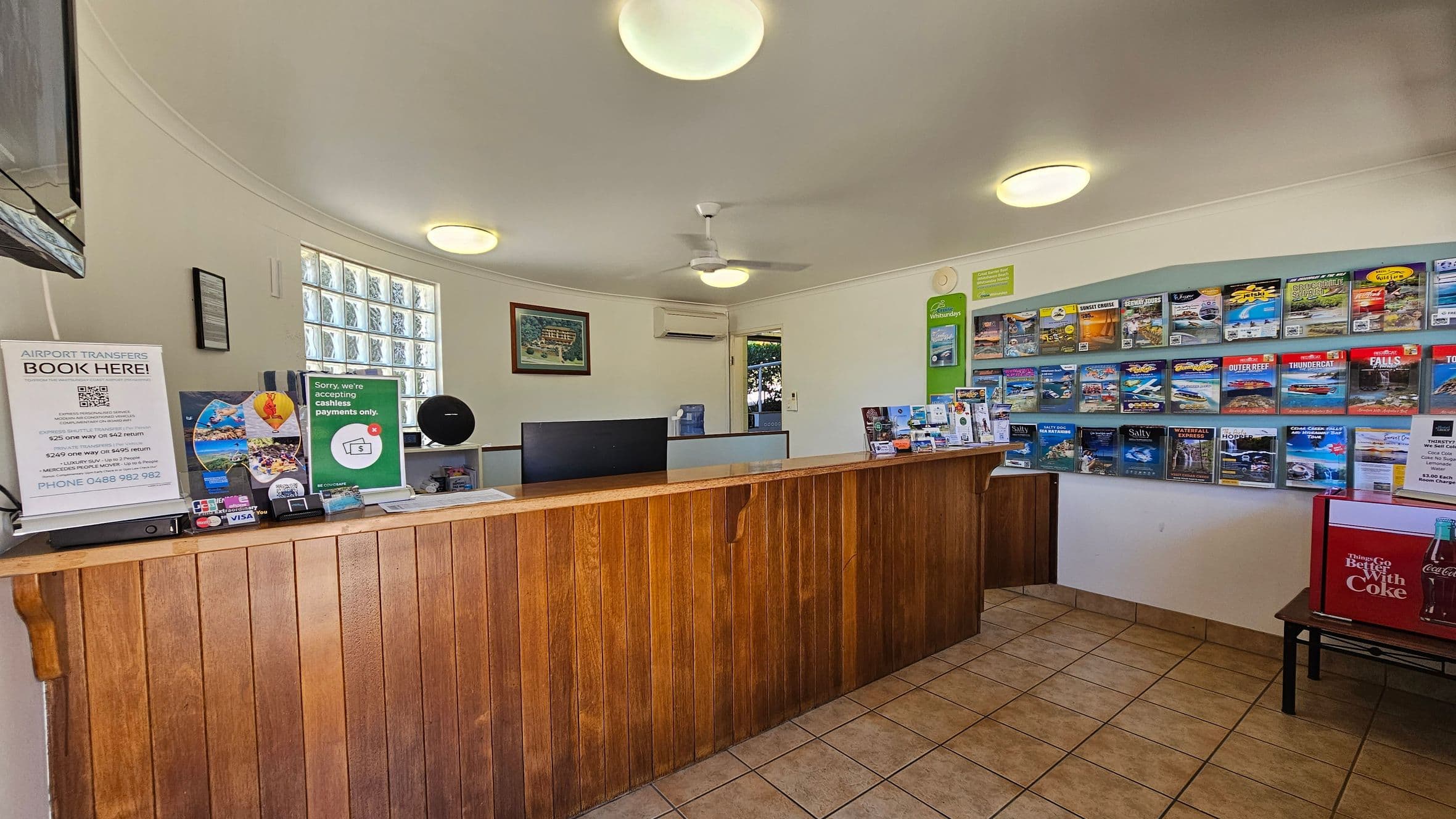 A reception area with a wooden counter in the foreground. On the left, there are signs for airport transfers and cashless payments. Behind the counter, a computer monitor and various brochures are visible. The right wall features a display of colourful brochures. The floor is tiled, and the ceiling has several round lights and a ceiling fan. A small Coca-Cola fridge is on the right side. A window with glass blocks is on the left wall, allowing natural light in.