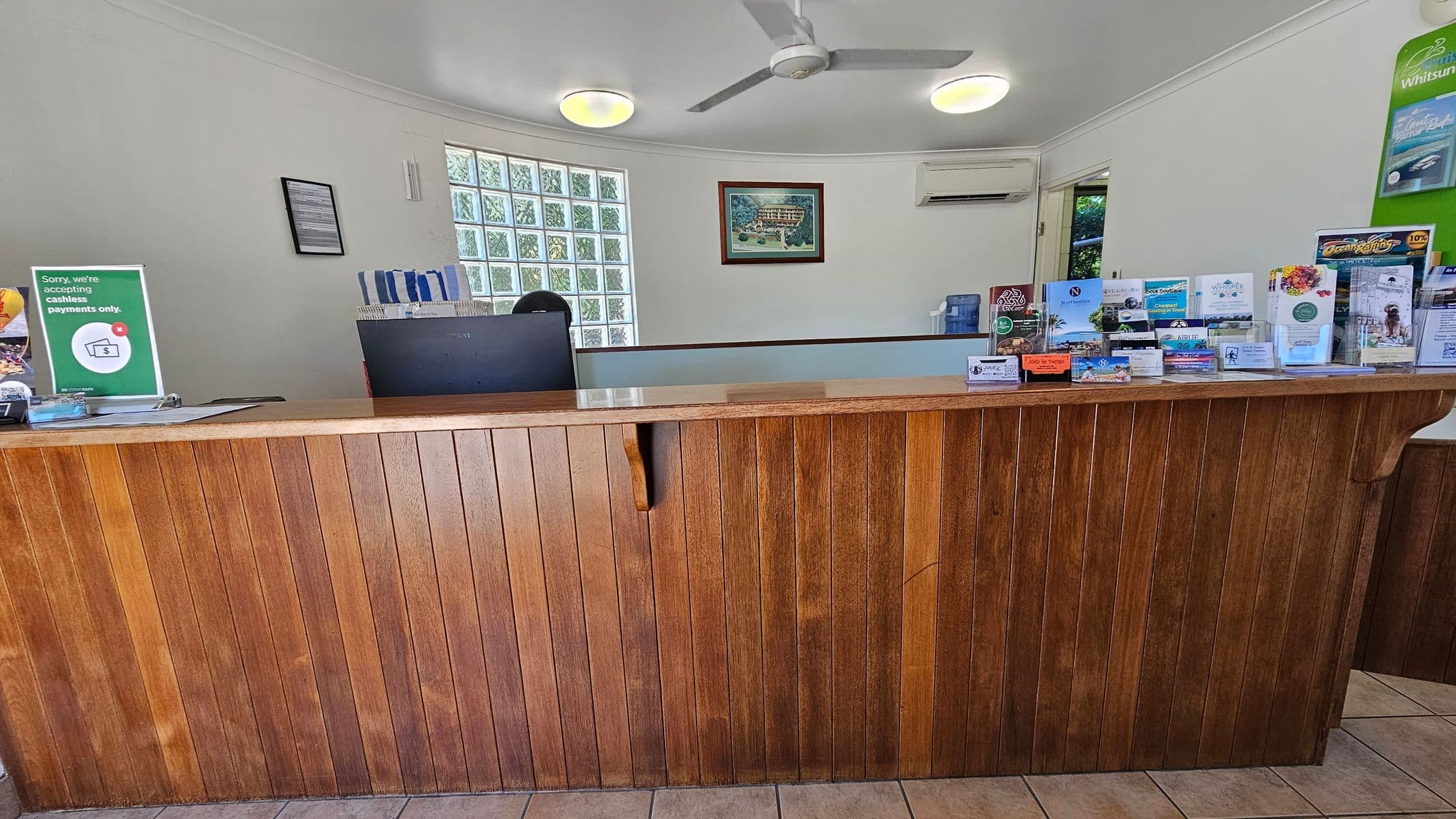 A reception area with a wooden counter in the foreground. On the right side of the counter, there are various brochures and business cards displayed. Behind the counter, a computer monitor is visible. The background features a wall with a framed picture, an air conditioning unit, and a glass block window. The ceiling has two round lights and a ceiling fan. The floor is tiled.