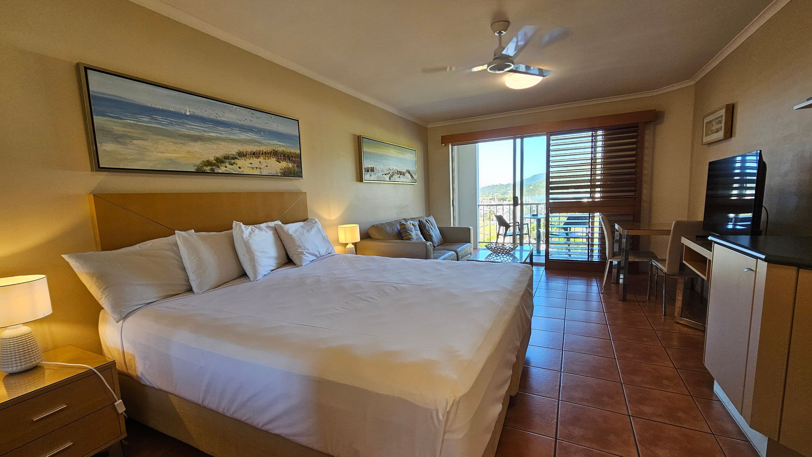 A hotel room with a large bed on the left, adorned with white pillows and sheets. A bedside table with a lamp is next to the bed. Above the bed, a landscape painting hangs on the wall. In the middle, there's a beige sofa with cushions and another painting above it. On the right, a flat-screen TV sits on a cabinet, with a small table and chair nearby. The room has a tiled floor and a ceiling fan. Sliding glass doors lead to a balcony with a view of hills and water.