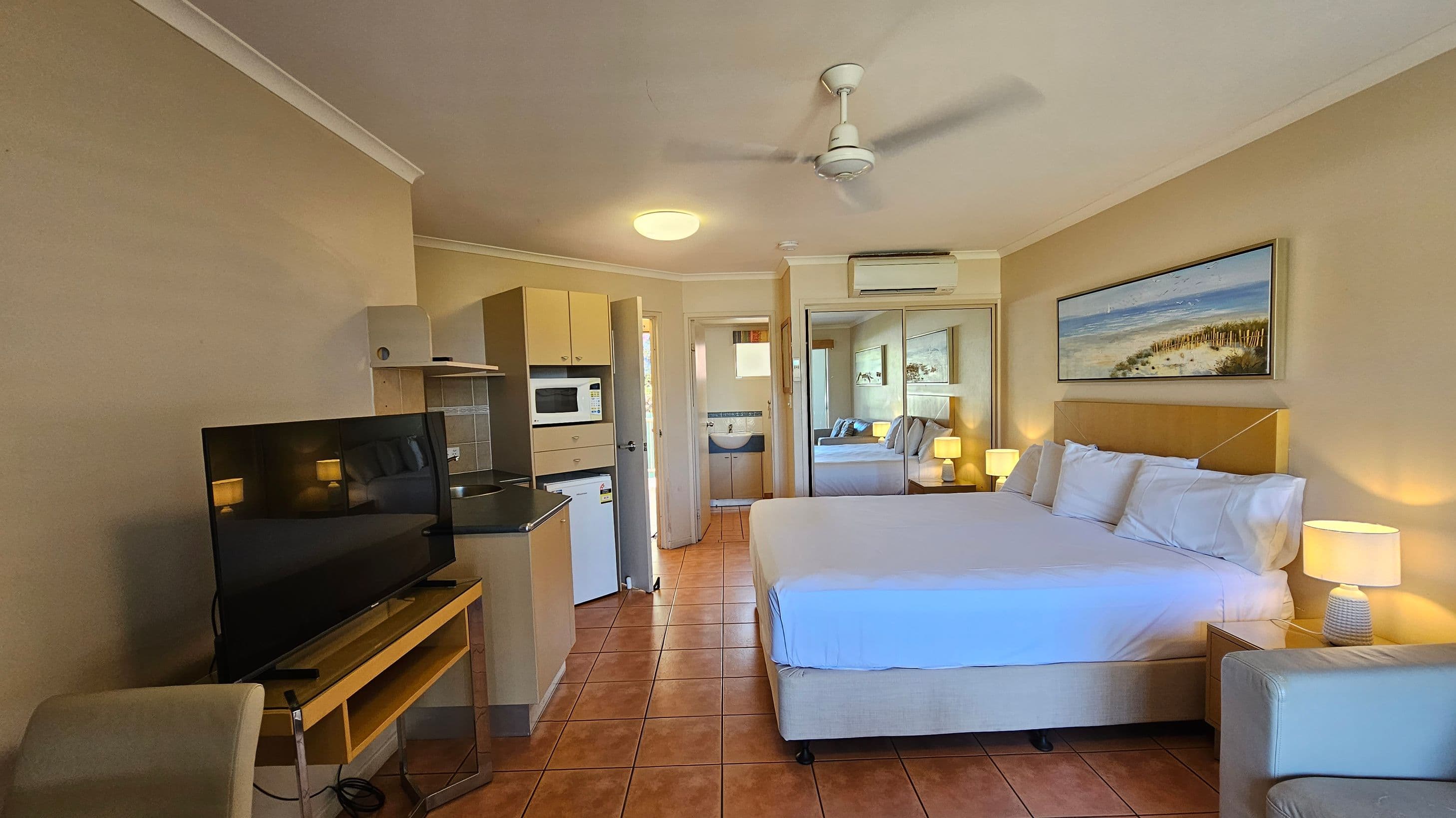 A hotel room with terracotta-tiled flooring and cream walls. On the right, there's a large bed with white linens, flanked by bedside tables with lamps. Above the bed is a framed beach painting. On the left, a flat-screen TV sits on a wooden stand. A kitchenette with a microwave, fridge, and sink is visible. In the background, a doorway leads to a bathroom. A ceiling fan and air conditioning unit are mounted above.