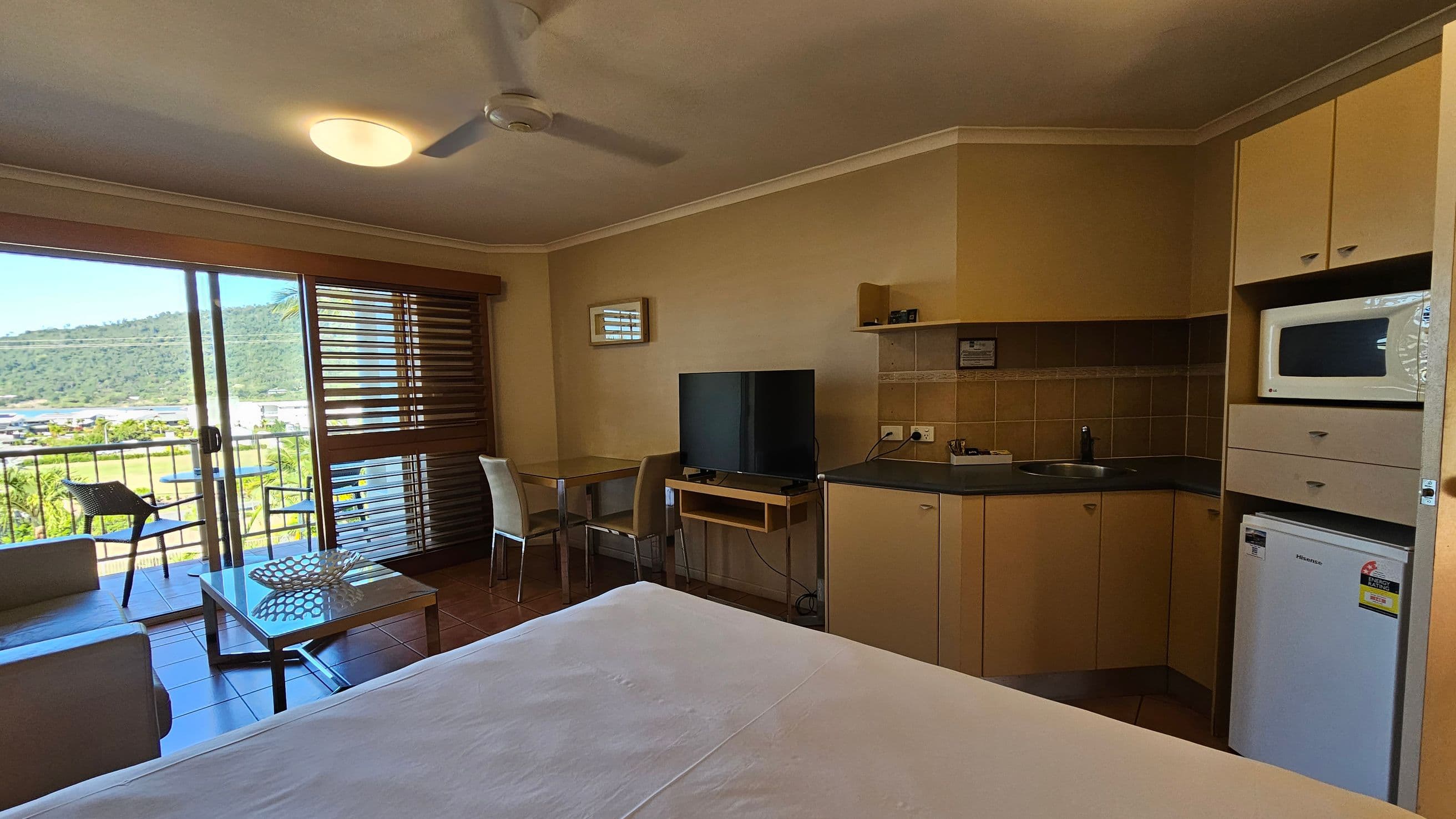 A room with a view of a balcony and hills in the background. On the left, there's a sliding glass door with wooden blinds, leading to a small balcony with a chair. Inside, a sofa and a coffee table are near the door. In the middle, a small dining table with two chairs is next to a flat-screen TV on a stand. On the right, a kitchenette with a microwave, sink, and mini-fridge. The ceiling has a fan and a light fixture, and the floor is tiled.