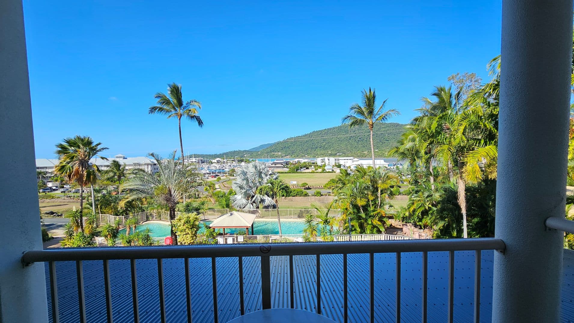 View from a balcony with a railing in the foreground. Below, a swimming pool is surrounded by palm trees and a small gazebo. In the middle distance, there are buildings and a marina with boats. A lush green hill rises in the background under a clear blue sky.