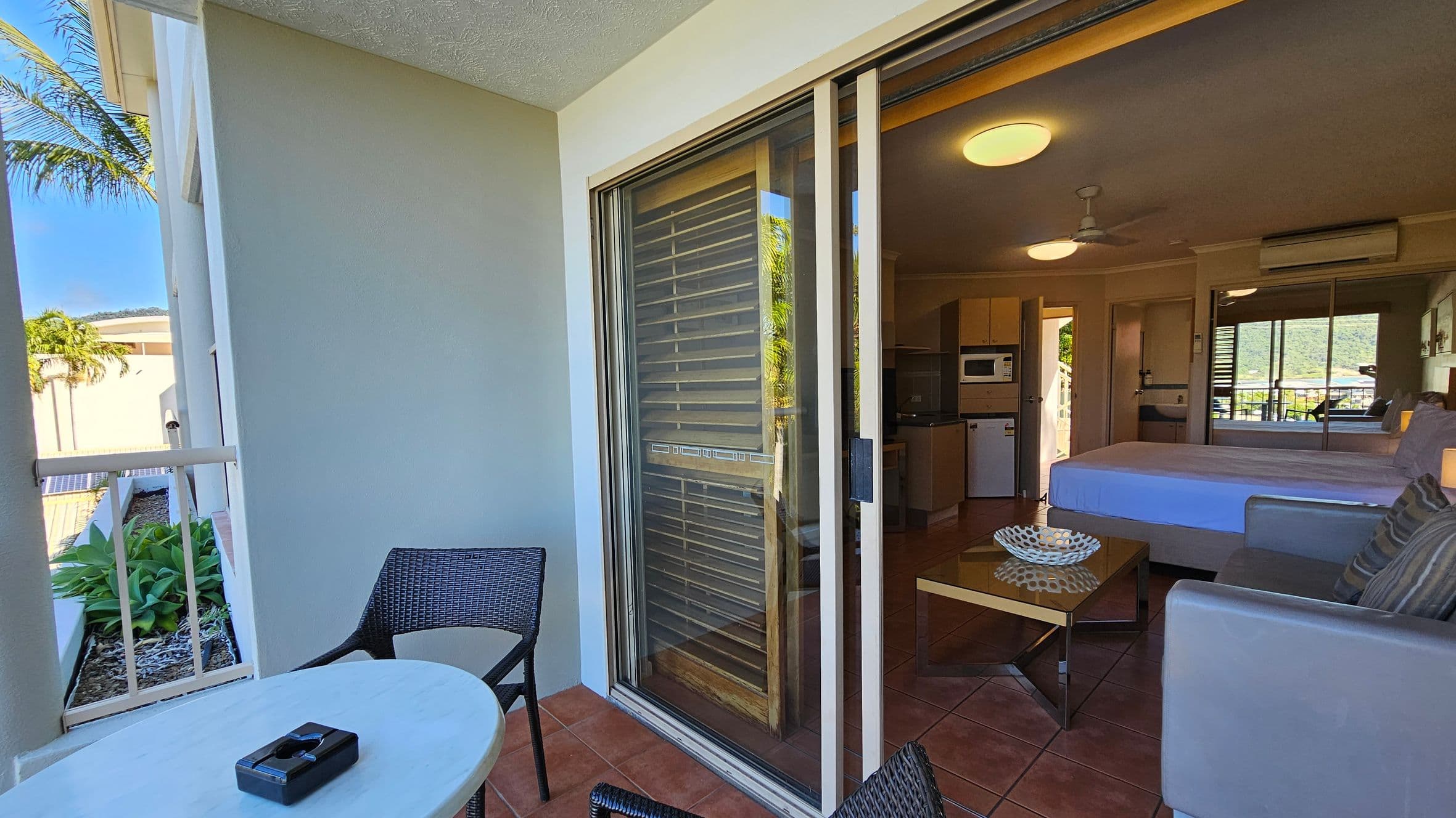 A balcony with terracotta tiles features a small round table and two wicker chairs. A sliding glass door leads to an interior room with a bed, a sofa, and a coffee table. The room has a ceiling fan and an air conditioning unit. A kitchenette with a microwave and a small fridge is visible. Outside, there are green plants and a partial view of a palm tree.