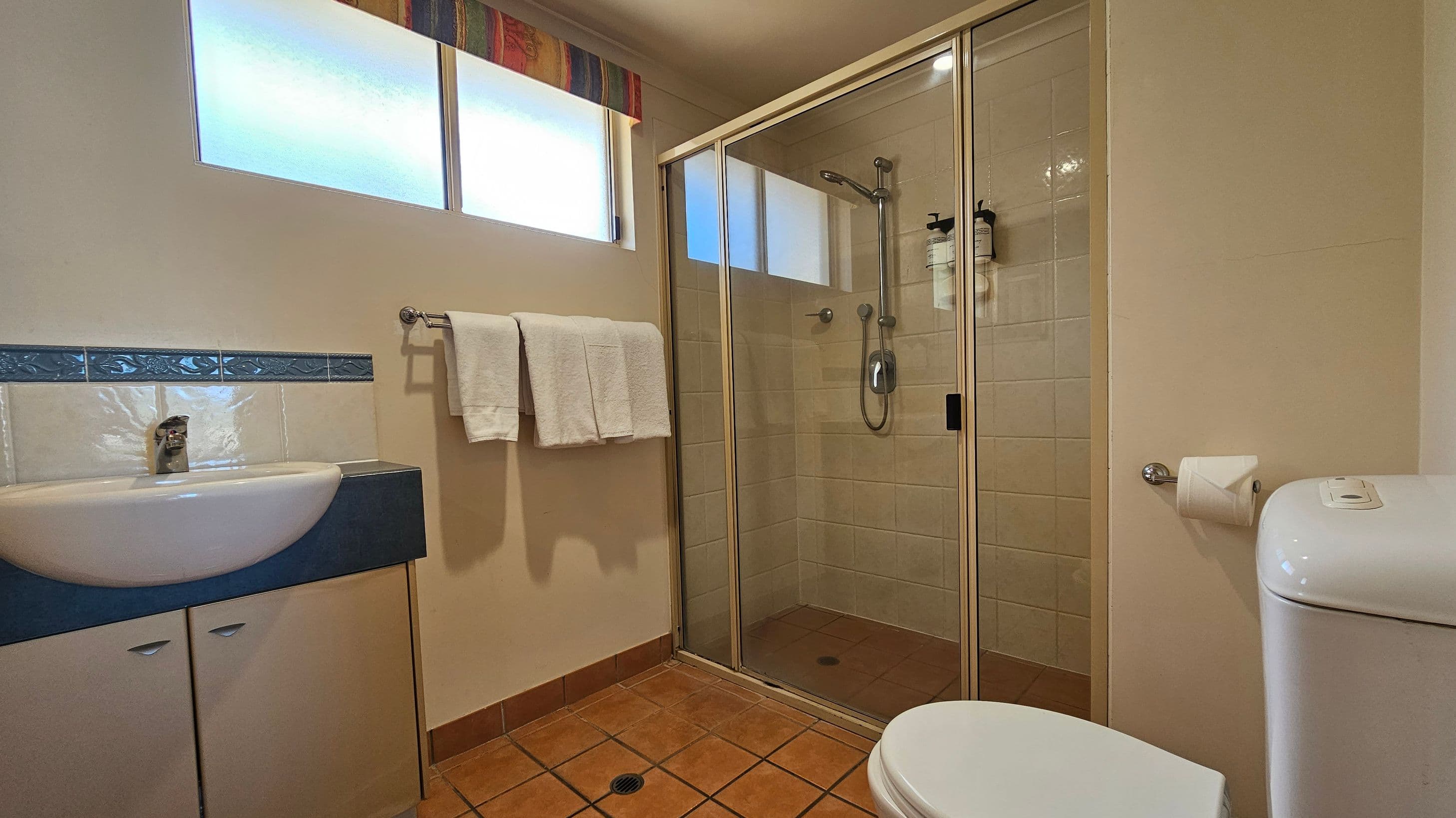 A bathroom with a tiled floor and cream walls. On the left, there's a sink with a blue countertop and a cabinet below, with a decorative blue tile border above. A towel rack with white towels is mounted on the wall. In the centre, a glass-enclosed shower with a sliding door and mounted dispensers. On the right, a toilet with a roll of toilet paper on a holder. Above, frosted windows with a colourful valance allow light in.