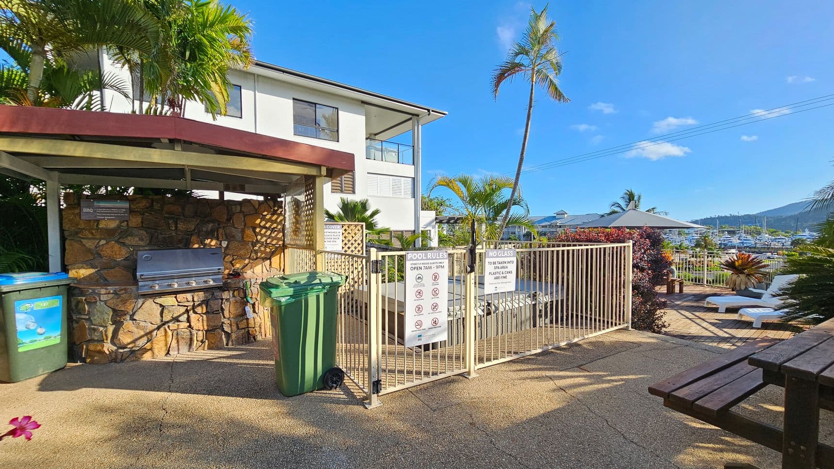 A pool area with a stone barbecue on the left, sheltered by a small roof. Two green bins are nearby. A white building with large windows is in the background. A gated entrance with pool rules is in the centre, leading to a deck with white sun loungers and a large umbrella. Palm trees and lush plants surround the area. In the distance, a marina with boats is visible, set against a backdrop of hills. A wooden bench is in the foreground on the right.