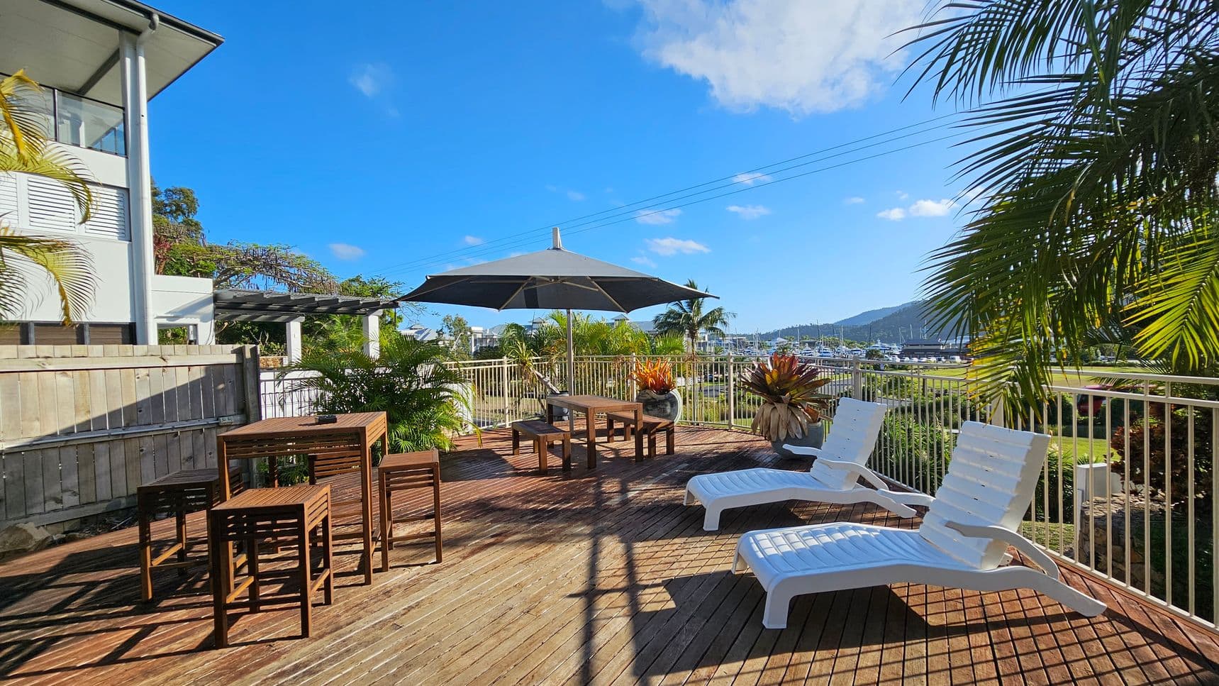 A wooden deck with two white sun loungers on the right and a wooden table with stools on the left. In the centre, a large black umbrella provides shade over a seating area. Potted plants are scattered around, and a metal railing encloses the space. In the background, there are views of a marina with boats and distant hills. A modern building with a wooden fence is on the left, and palm fronds partially frame the scene.