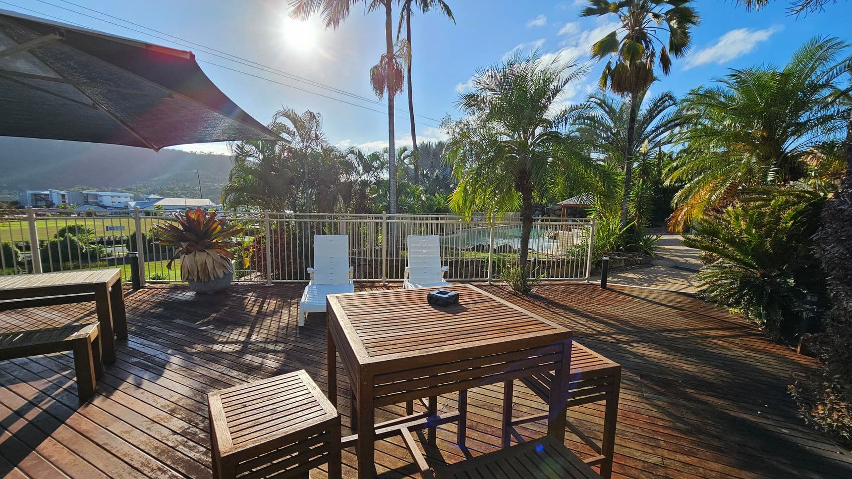 Wooden deck with a table and benches in the foreground. Two white sun loungers are positioned near a metal railing. A large potted plant is on the left. Palm trees and lush greenery are in the background, with a glimpse of a pool area. A sunshade partially covers the deck on the left. The sun is shining brightly, casting shadows on the deck.