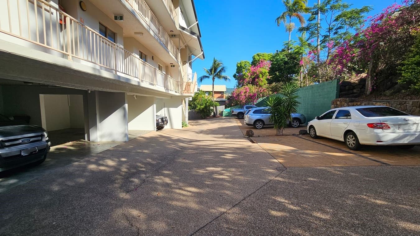 A parking area with several cars is visible. On the left, a building with a light-coloured exterior and a balcony runs along the side. Below the balcony, there are covered parking spaces with cars parked. On the right, a white car is parked near a green fence, with flowering bushes and trees behind it. The ground is paved, and shadows from the trees create patterns on the surface. A palm tree is visible in the background.