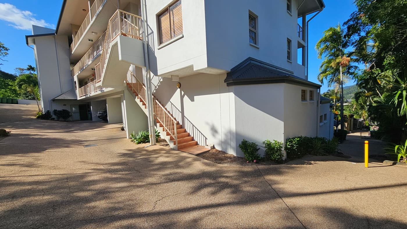 A multi-storey building with white walls and a grey roof is on the right. A staircase with a metal railing leads up to the first floor. The building has several windows with shutters. On the left, there is a driveway with a parked car under a covered area. The ground is paved, and there are small plants and shrubs along the building's base. In the background, there are palm trees and a view of distant hills. Shadows of trees are cast on the ground.