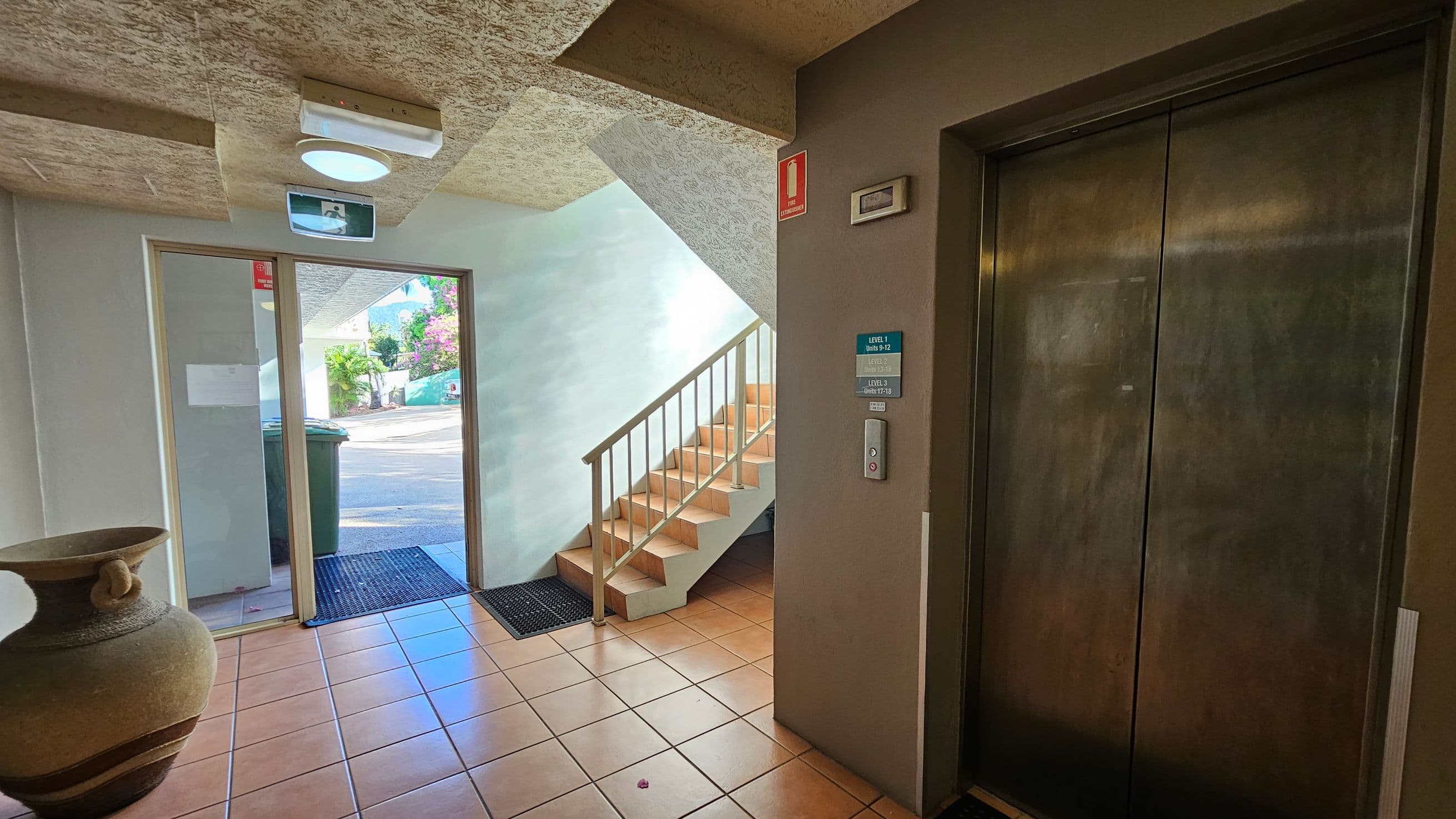 A hallway with a tiled floor and textured ceiling. On the right, there is a closed lift with a control panel and signage indicating floor levels. In the centre, a staircase with a metal railing leads upwards. On the left, a large decorative pot sits near a glass door, which opens to the outside. A green wheelie bin is visible through the door. A light fixture and an exit sign are mounted on the ceiling.