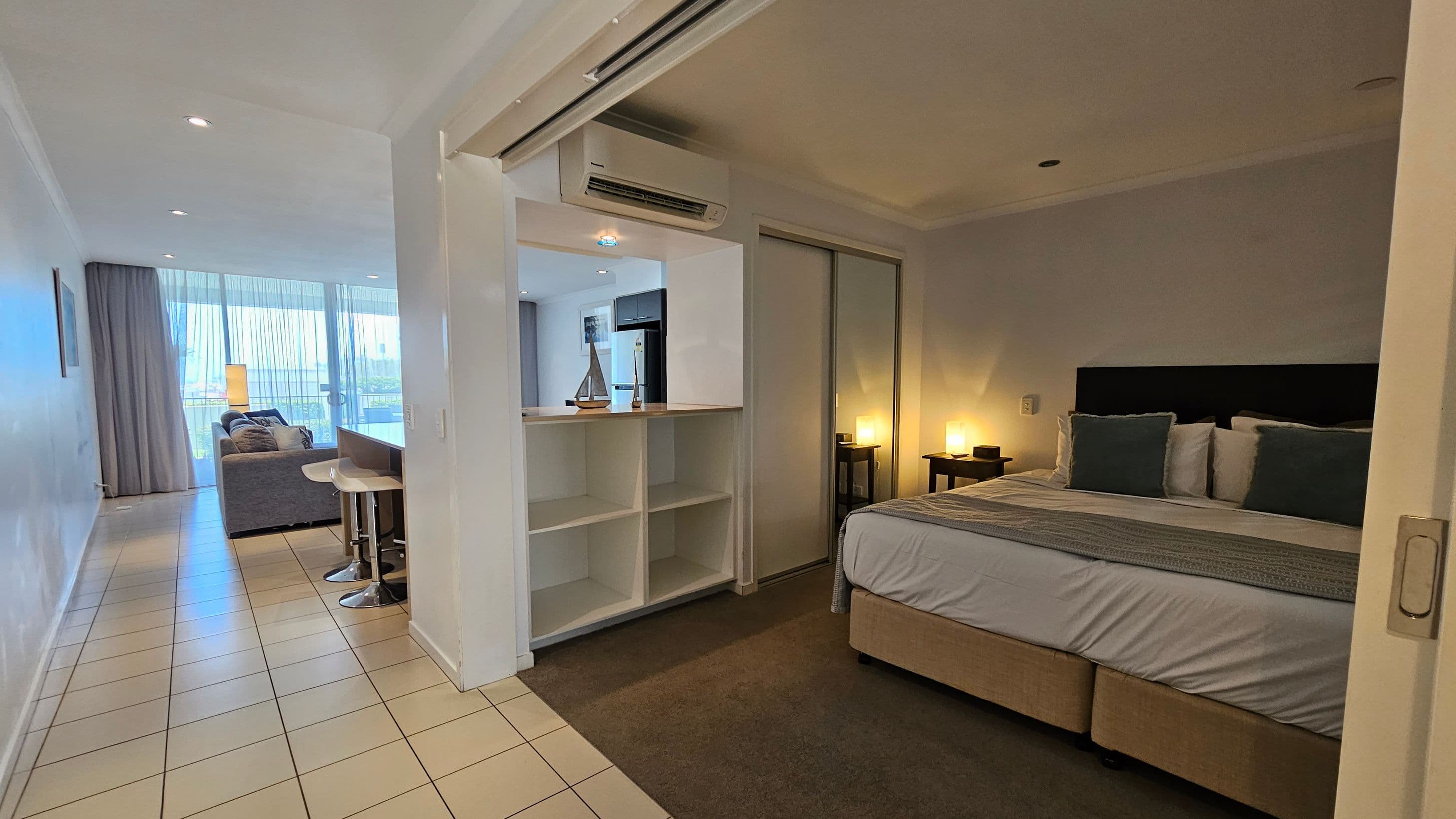 A modern hotel room with a tiled floor. On the right, there's a bedroom with a double bed, adorned with blue and white pillows, and bedside tables with lamps. In the centre, a partition with shelves separates the bedroom from the living area. The left side features a living space with a grey sofa, a small table, and a large window with sheer curtains. The ceiling has recessed lighting, and an air conditioning unit is mounted above the partition.