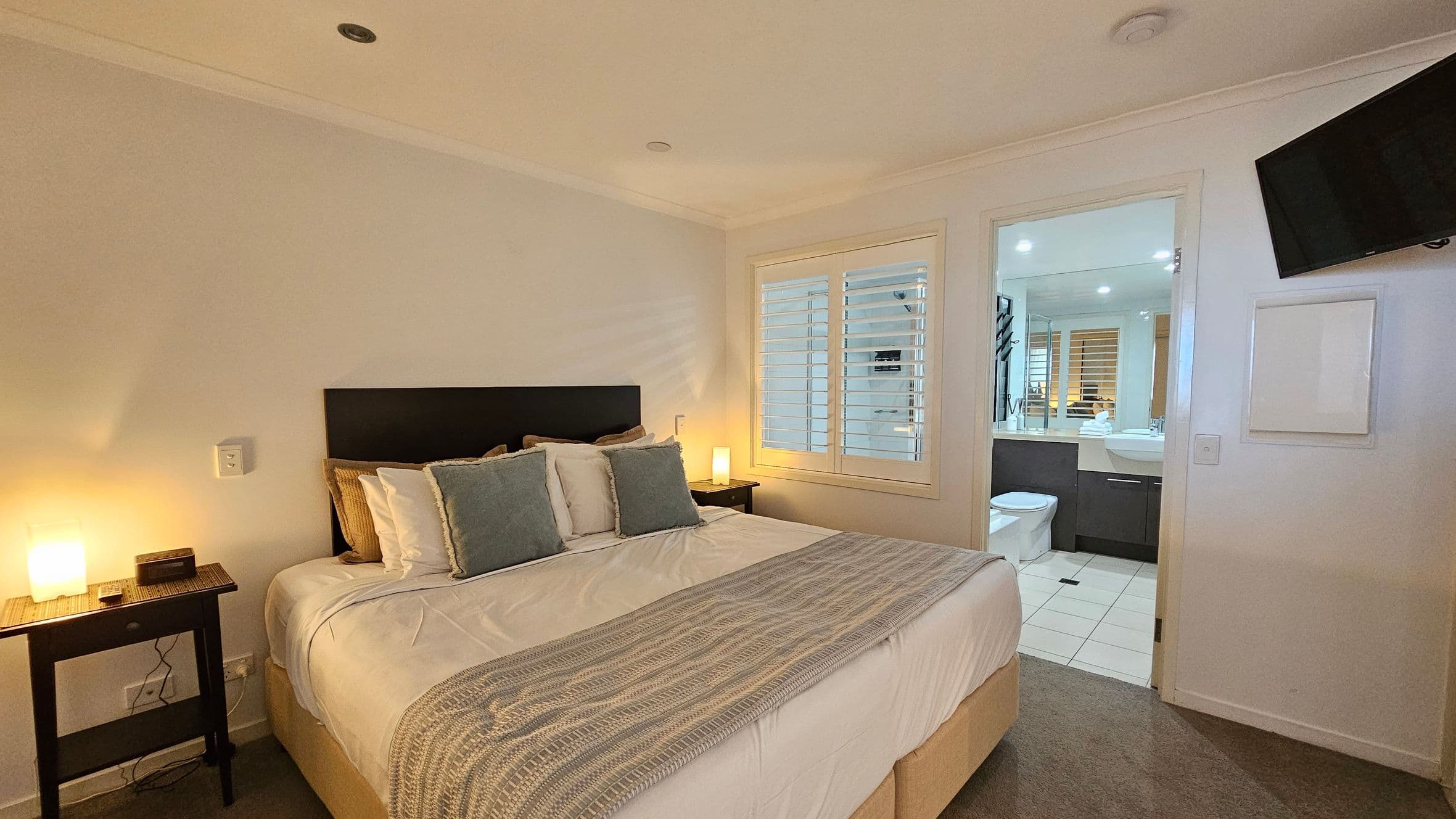 A bedroom with a large bed in the centre, featuring a dark headboard and multiple pillows. On either side of the bed are small bedside tables with lamps. To the right, there's a window with white shutters and an open door leading to a bathroom. The bathroom has a toilet and a sink with a mirror. A wall-mounted TV is on the right wall. The floor is carpeted, and the ceiling has recessed lighting.