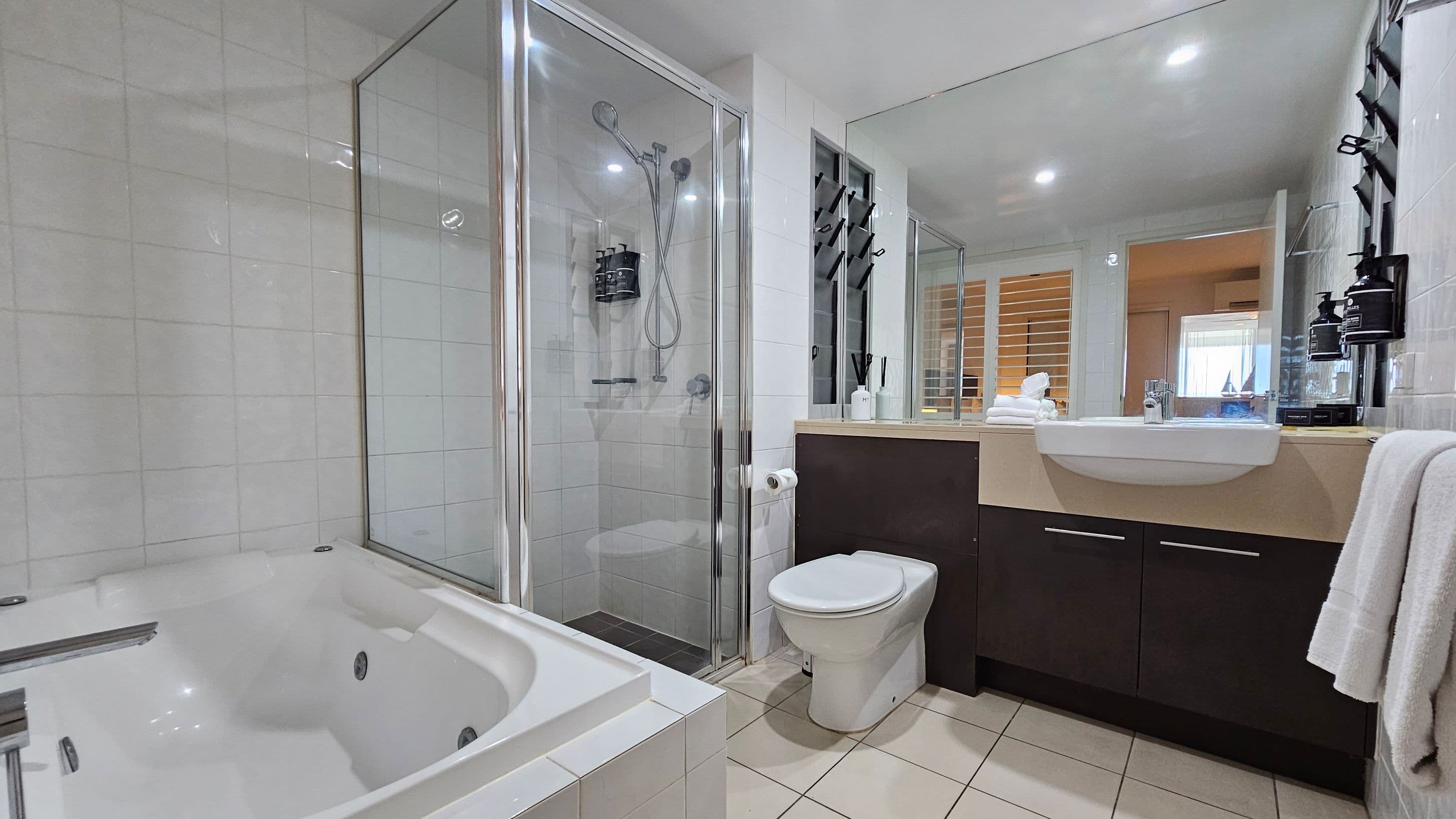 A modern bathroom with a white tiled floor. On the left, there's a large white bathtub. In the centre, a glass-enclosed shower with a chrome showerhead and dispensers. To the right, a white toilet next to a dark vanity with a white sink. Above the sink, a large mirror reflects the room. A towel hangs on the right side, and toiletries are neatly arranged on the counter. The ceiling has recessed lighting.