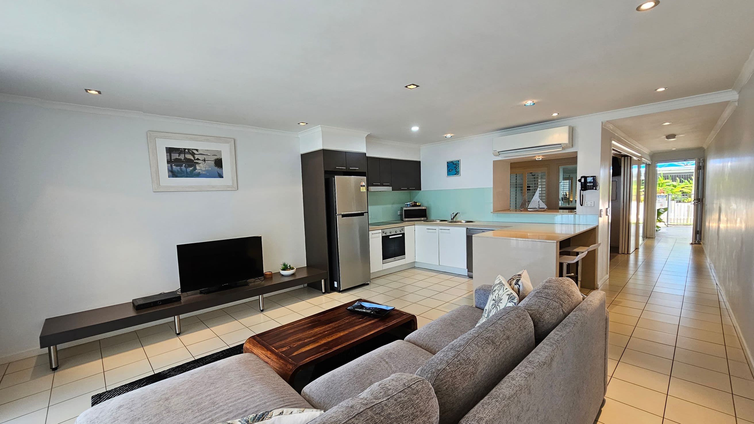 Open-plan living area with a grey sofa and wooden coffee table in the foreground. A flat-screen TV sits on a low black stand against the left wall, beneath a framed picture. The kitchen on the right features a stainless steel fridge, oven, and white cabinetry. A breakfast bar with stools is adjacent. The hallway leads to a door with a view of greenery outside. The floor is tiled, and recessed lighting is in the ceiling.