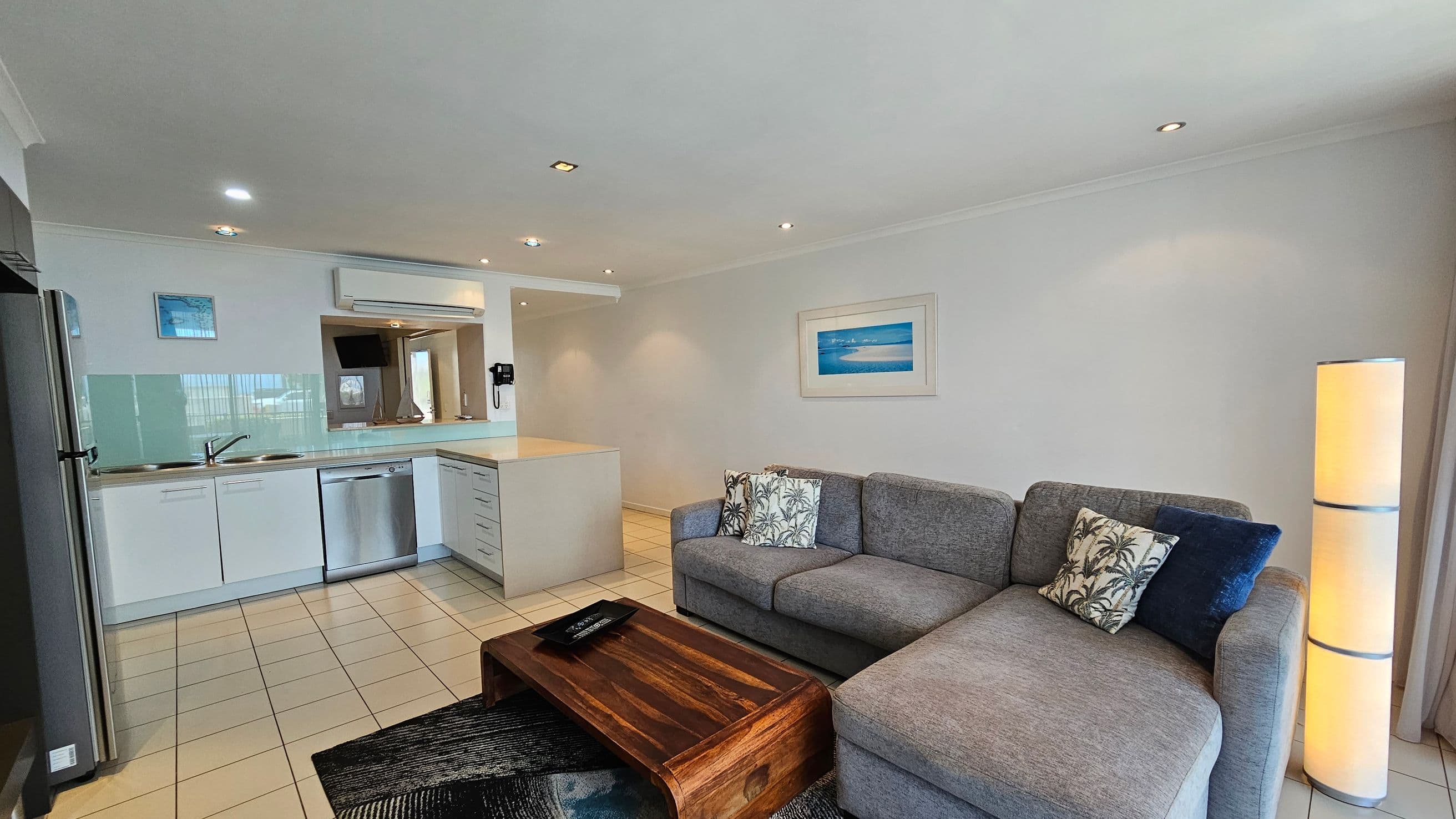 A modern living area with an open-plan kitchen on the left, featuring white cabinets, a stainless steel dishwasher, and a sink. The floor is tiled. On the right, a grey L-shaped sofa with patterned cushions sits next to a tall, cylindrical lamp. A wooden coffee table is in front of the sofa. A framed picture hangs on the wall above the sofa. The ceiling has recessed lighting.