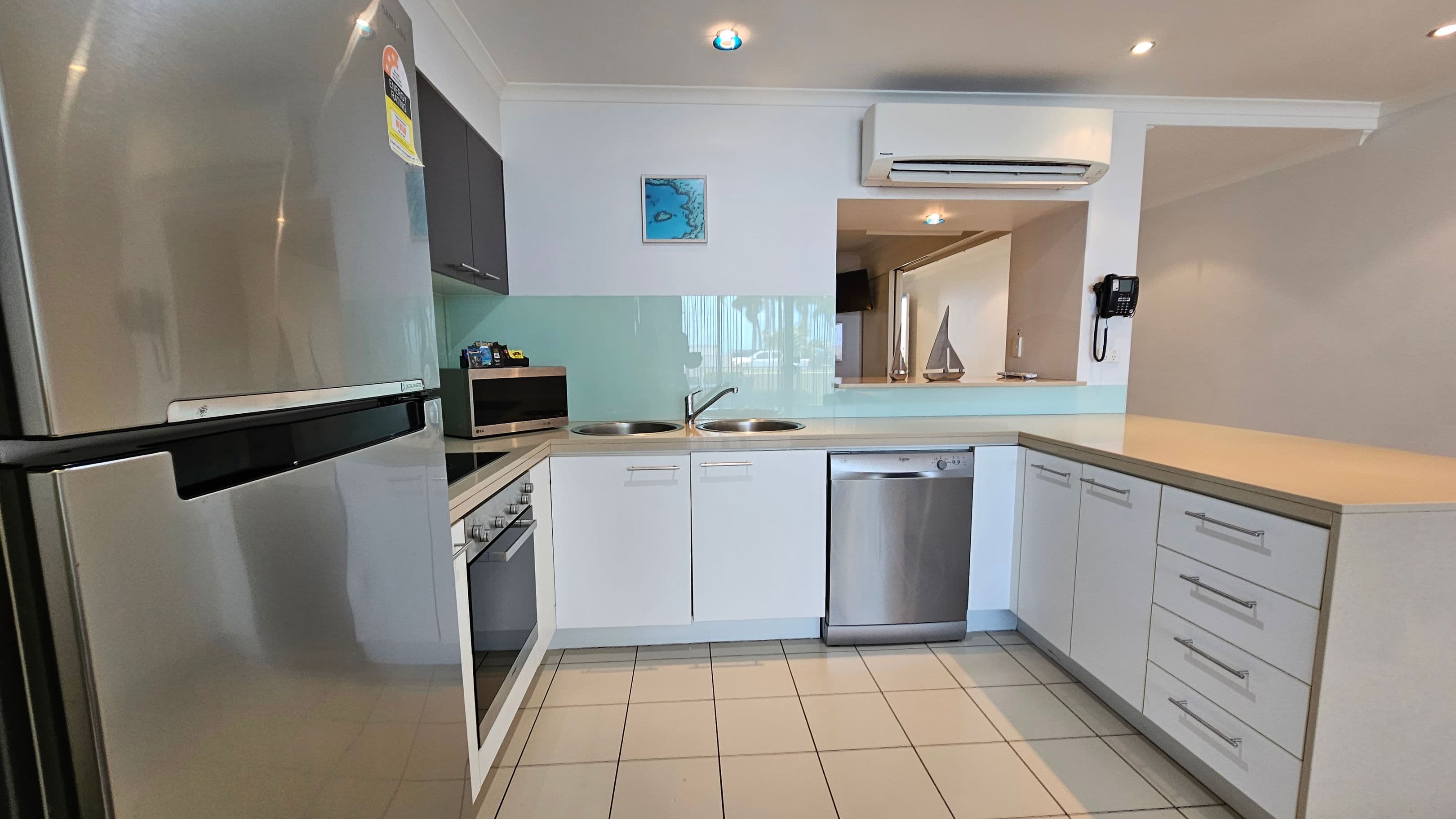 Modern kitchen with a stainless steel fridge on the left, adjacent to a microwave and an oven. In the centre, there are two sinks with a light green splashback. Below the sinks is a dishwasher. On the right, a counter with multiple drawers. Above, a wall-mounted air conditioner and a small framed picture. A phone is mounted on the wall. The floor is tiled, and the ceiling has recessed lighting. A small decorative sailboat is visible through an opening in the wall.