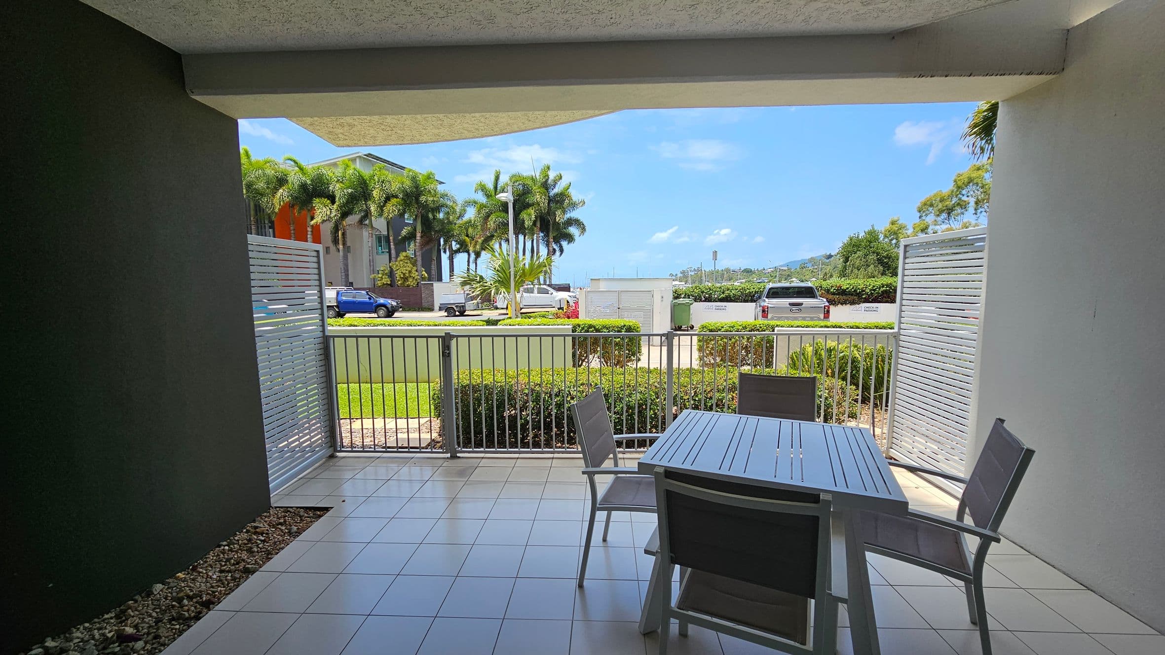 A covered patio with a tiled floor features a table and chairs. The patio is enclosed by a metal railing and has louvred panels on either side. Beyond the patio, there is a grassy area with neatly trimmed hedges. In the background, a road with parked vehicles, including a blue truck, is visible. Tall palm trees and a modern building are on the left, with a view of distant hills on the right.