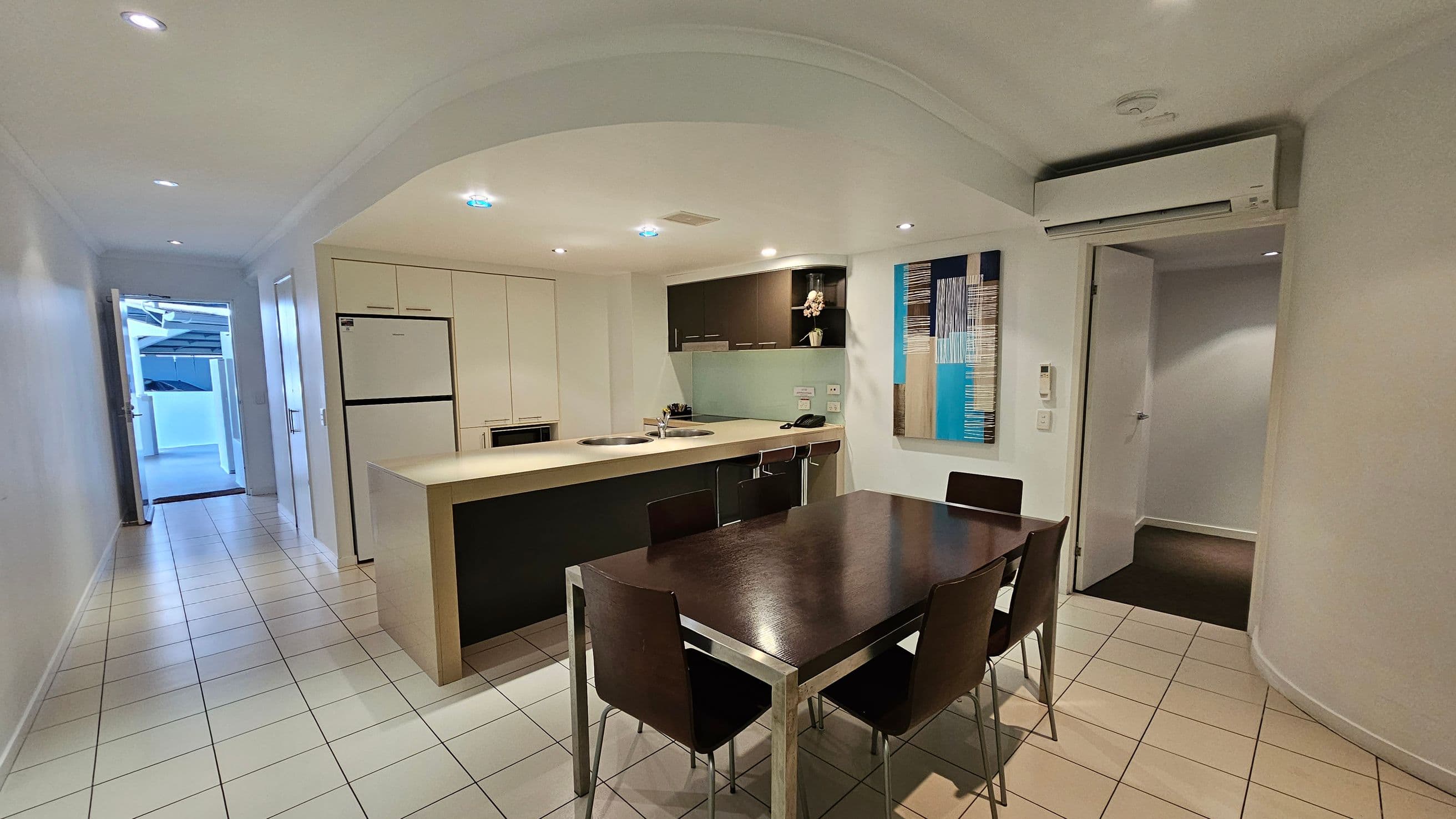 A modern kitchen and dining area with a tiled floor. On the left, there's a hallway leading to an open door. The kitchen features a large fridge, a microwave, and a counter with a sink. Above the counter are dark cabinets and a decorative vase. A colourful abstract painting is on the wall. The dining area has a dark wooden table with four chairs. An air conditioning unit is mounted on the wall to the right, near a partially open door. The ceiling has recessed lighting.