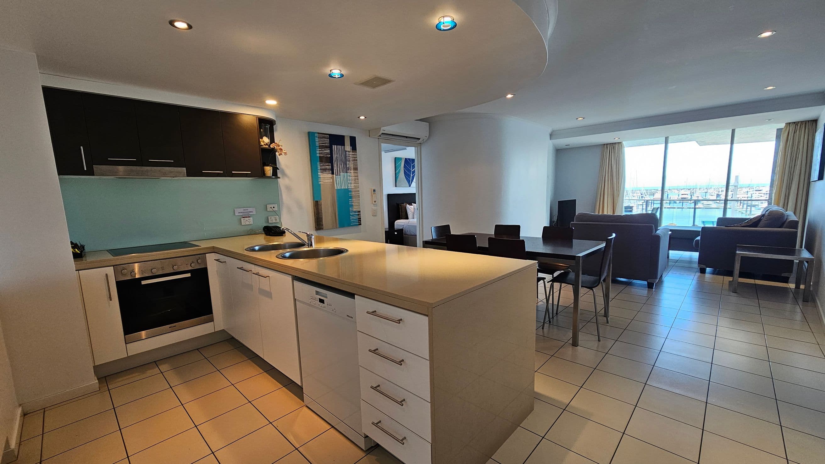 Modern open-plan kitchen and living area. On the left, a kitchen with black upper cabinets, a light-coloured countertop, and built-in appliances. In the centre, a dining table with black chairs. On the right, a living room with two sofas facing a large window with a view of a marina. The floor is tiled, and the ceiling has recessed lighting.
